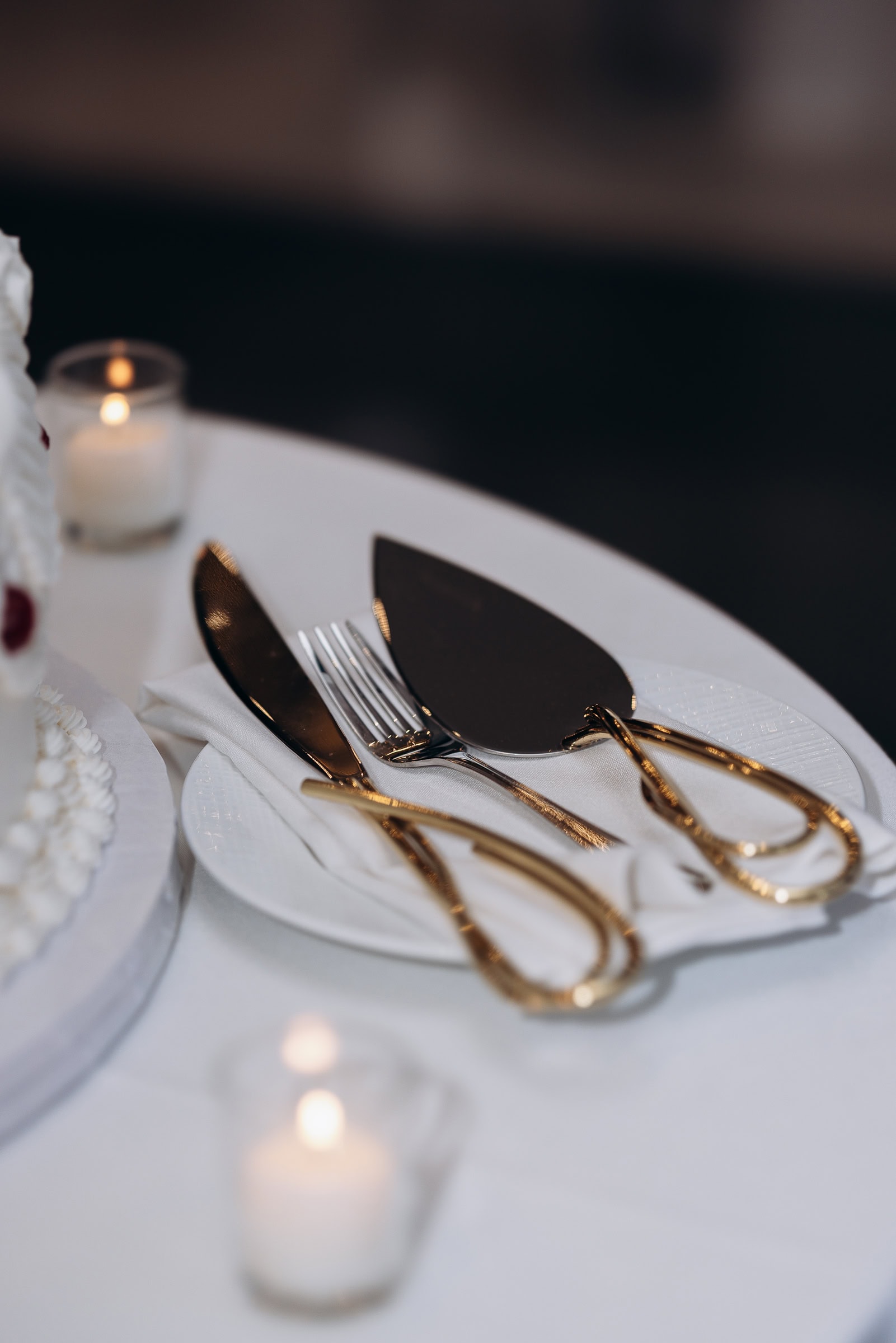 Close-up of gold-handled cake knife and server on white plate with napkin, next to a white wedding cake and lit candle at Hudson House, Jersey City.