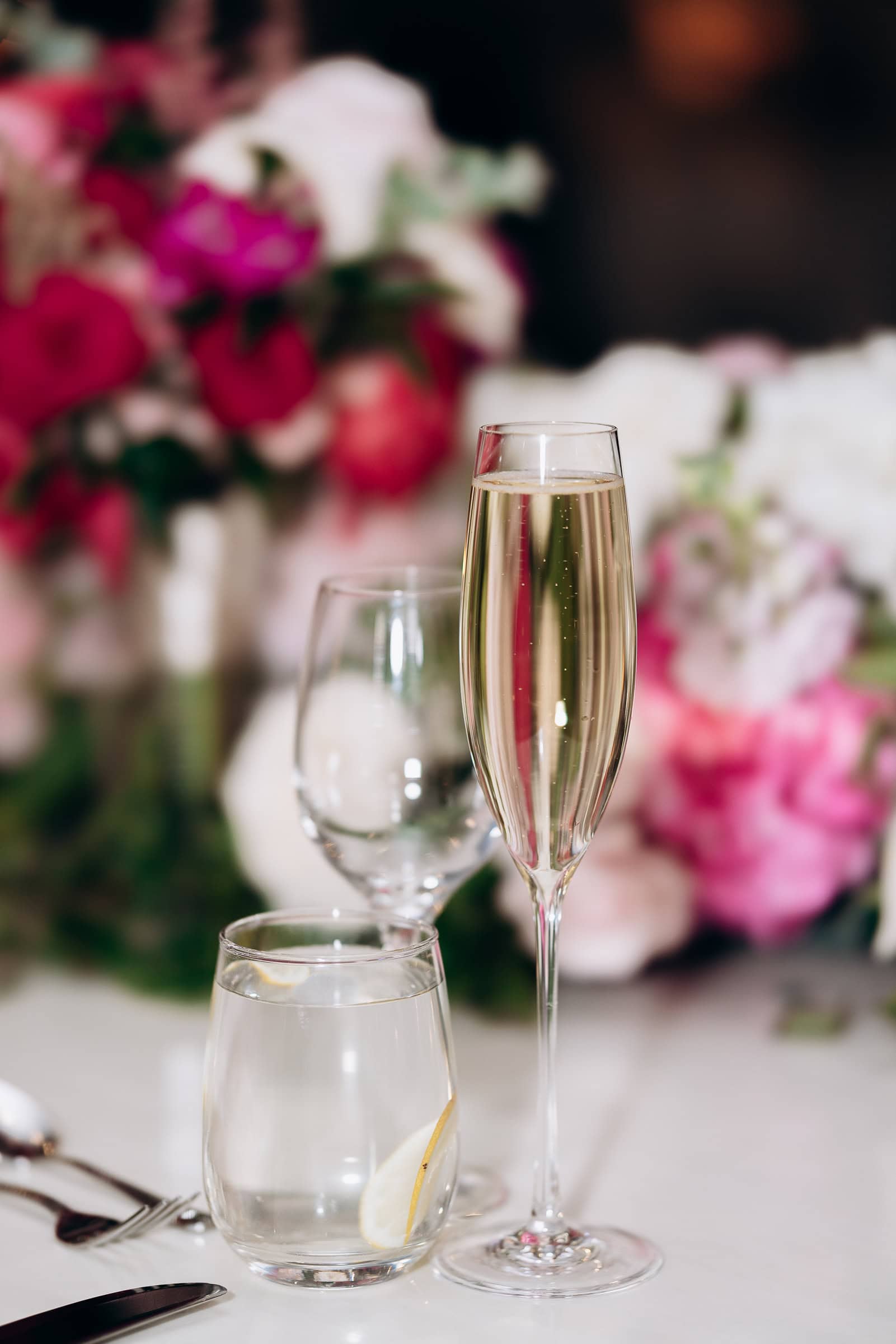 Close-up of a champagne flute filled with bubbly champagne on a table with a water glass and blurred pink and white flowers in the background at Hudson House, Jersey City.