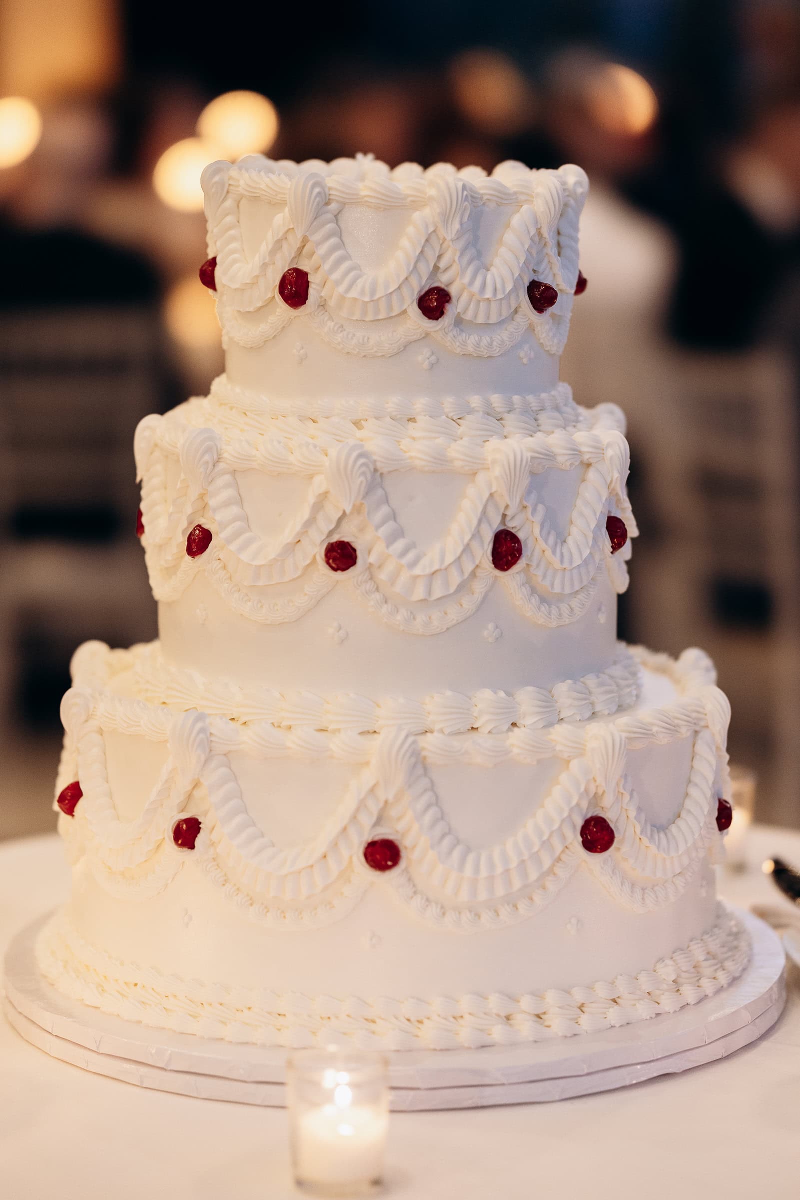 Close-up of a multi-tiered white wedding cake with intricate piped frosting and red decorative accents, placed on a white table with a lit candle nearby.