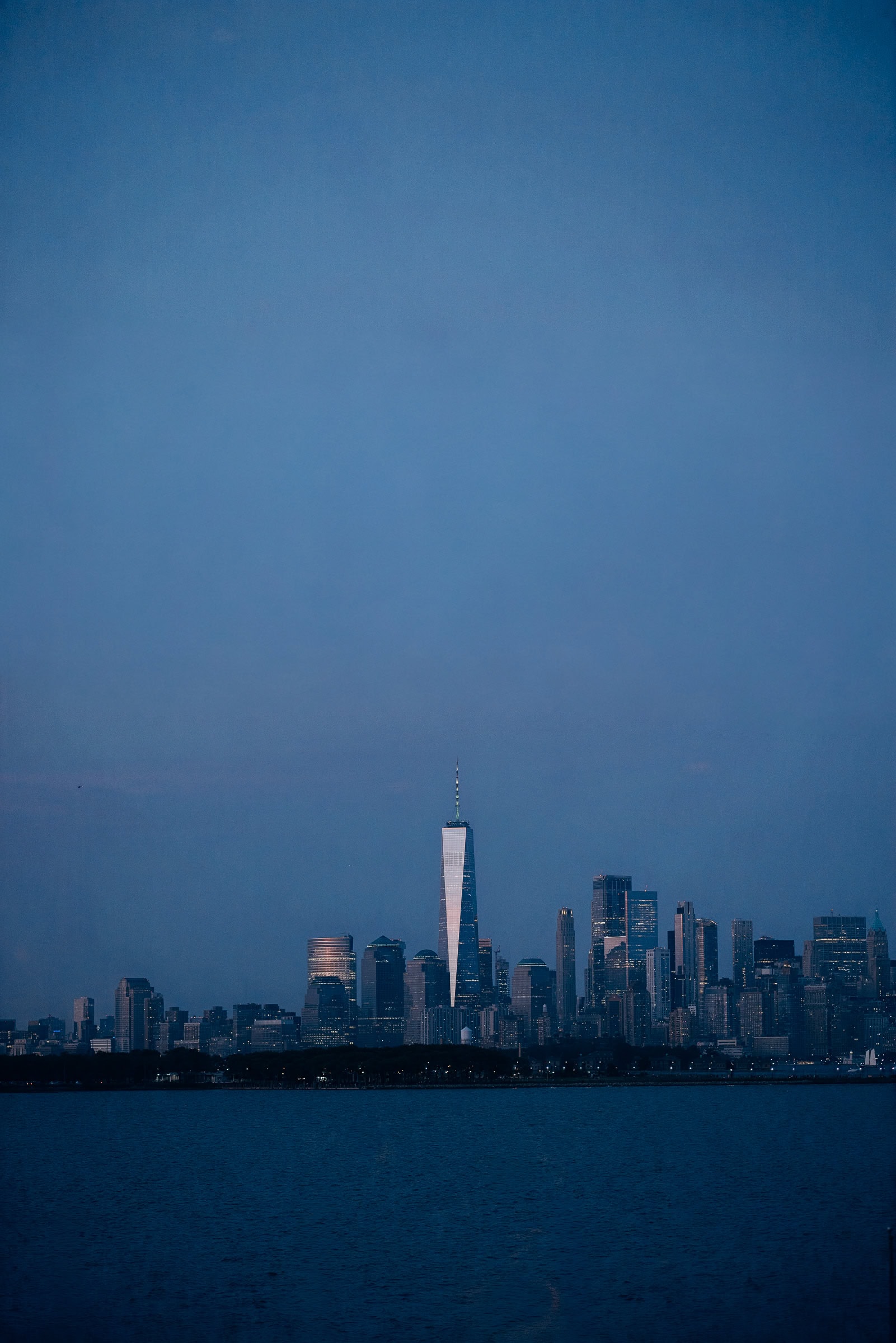 Manhattan skyline at twilight with One World Trade Center illuminated, viewed from across the water with reflections on glass.