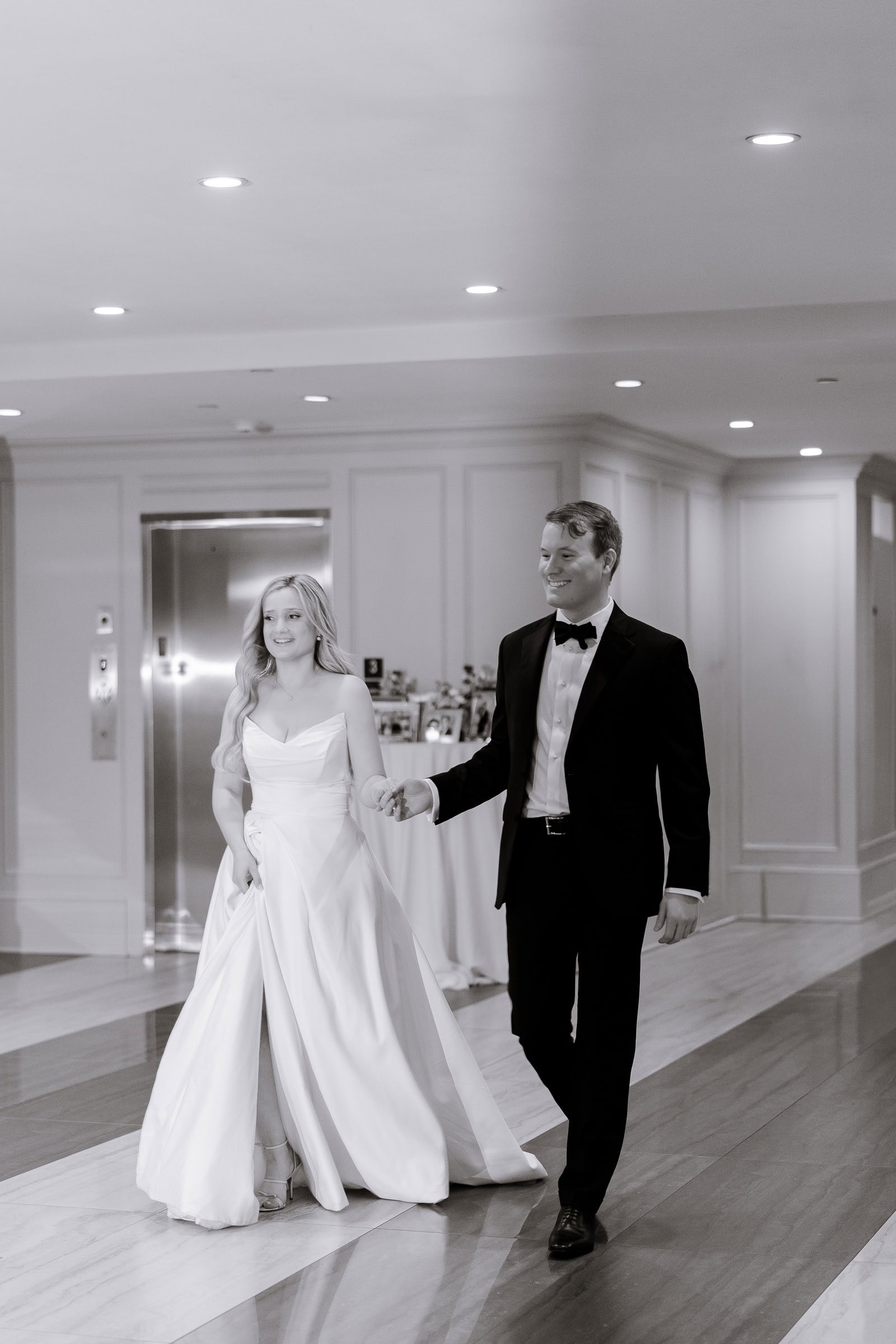 Bride in satin wedding dress with thigh-high slit and groom in black tuxedo holding hands walking indoors at Hudson House, Jersey City, NJ.