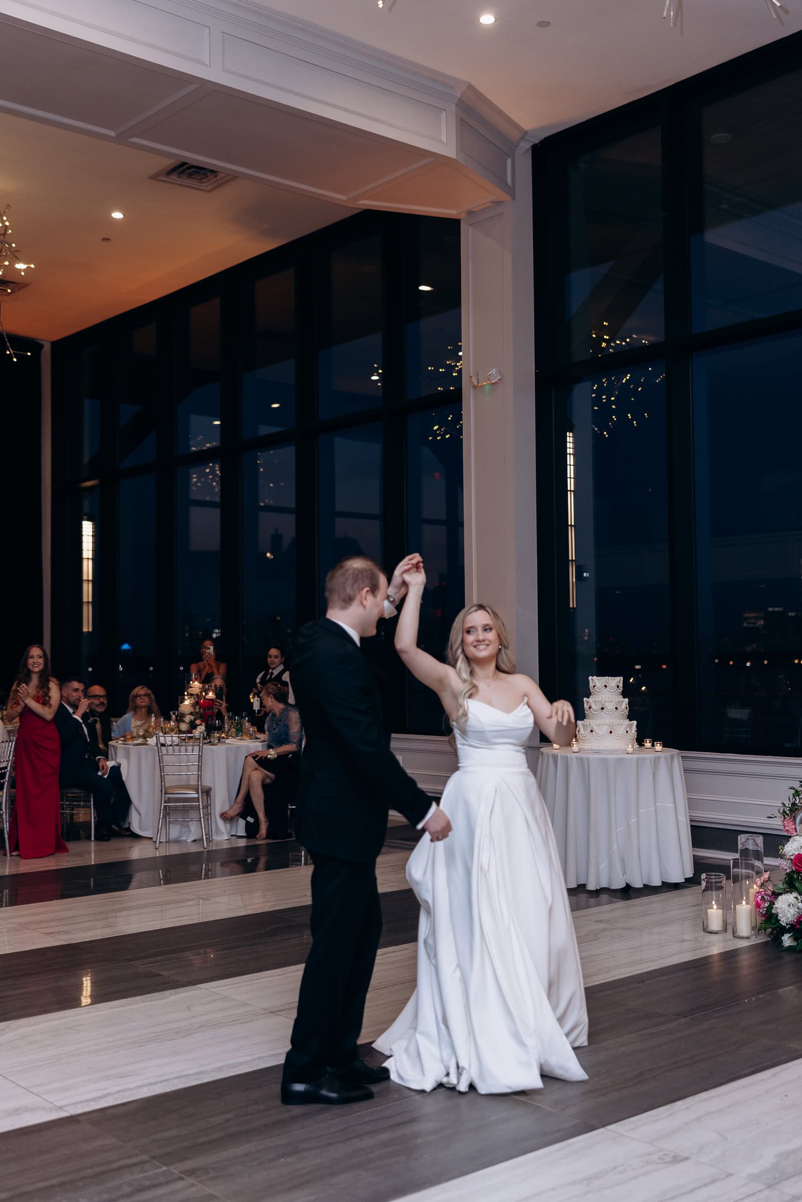 Bride in white gown and groom in black suit dancing at Hudson House wedding reception with city skyline visible through large windows.