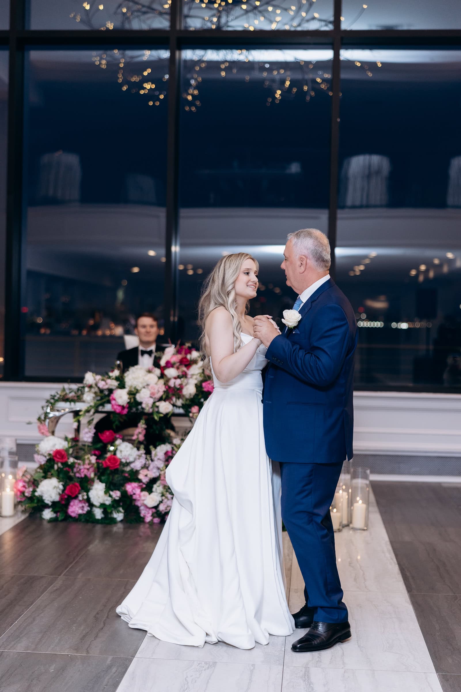 Bride in white gown dancing with her father in navy suit at Hudson House, Jersey City, with floral arrangements and candles in background.