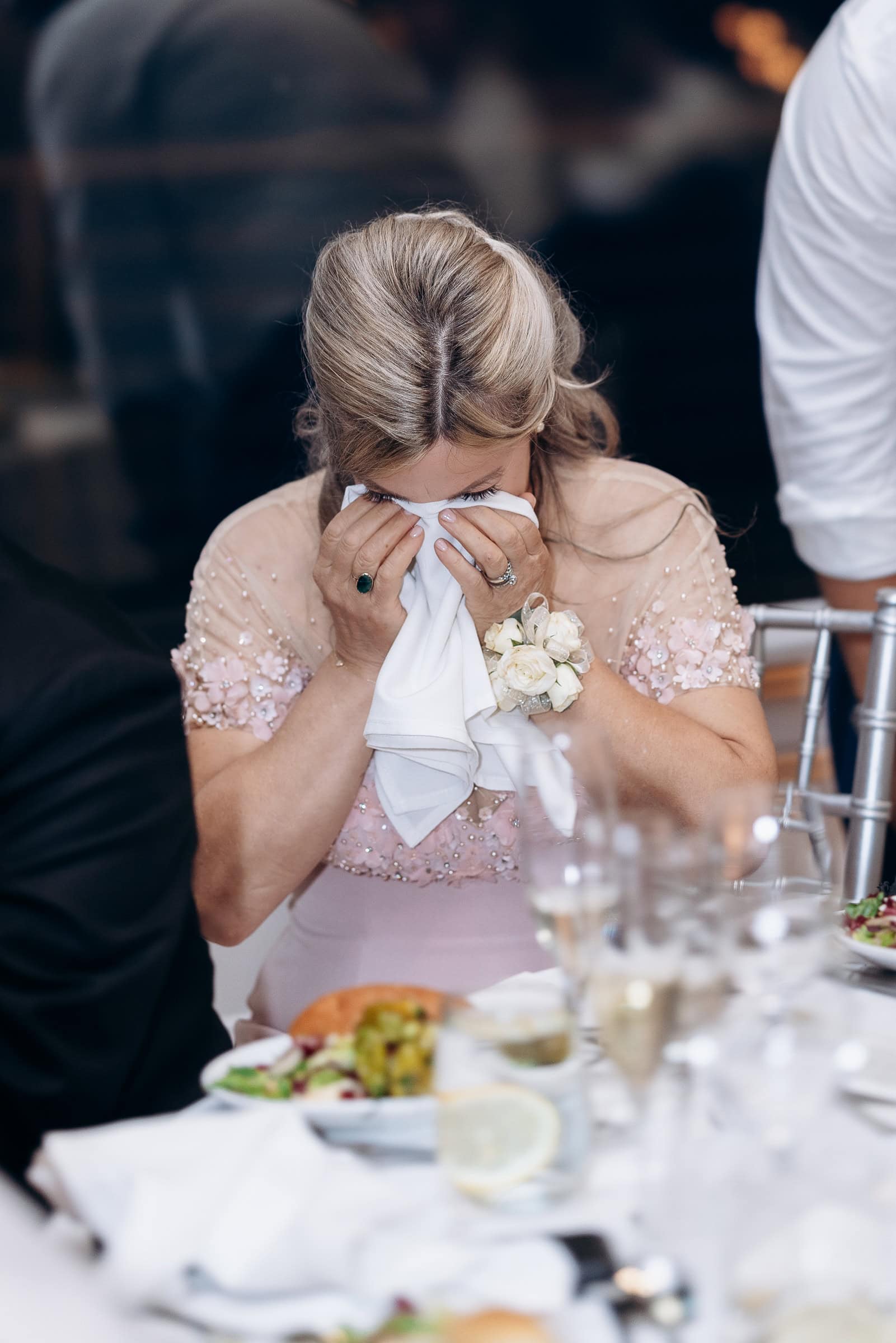 Woman in blush dress wiping tears with napkin, man in white shirt leaning in support, seated at a wedding reception table with plates and glasses.