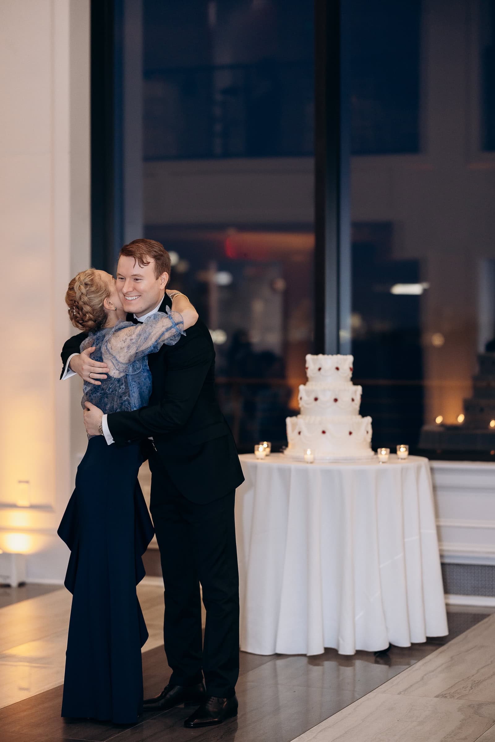 Two guests in formal attire sharing an intimate embrace near a tiered wedding cake at Hudson House, Jersey City, NJ, during a wedding reception.