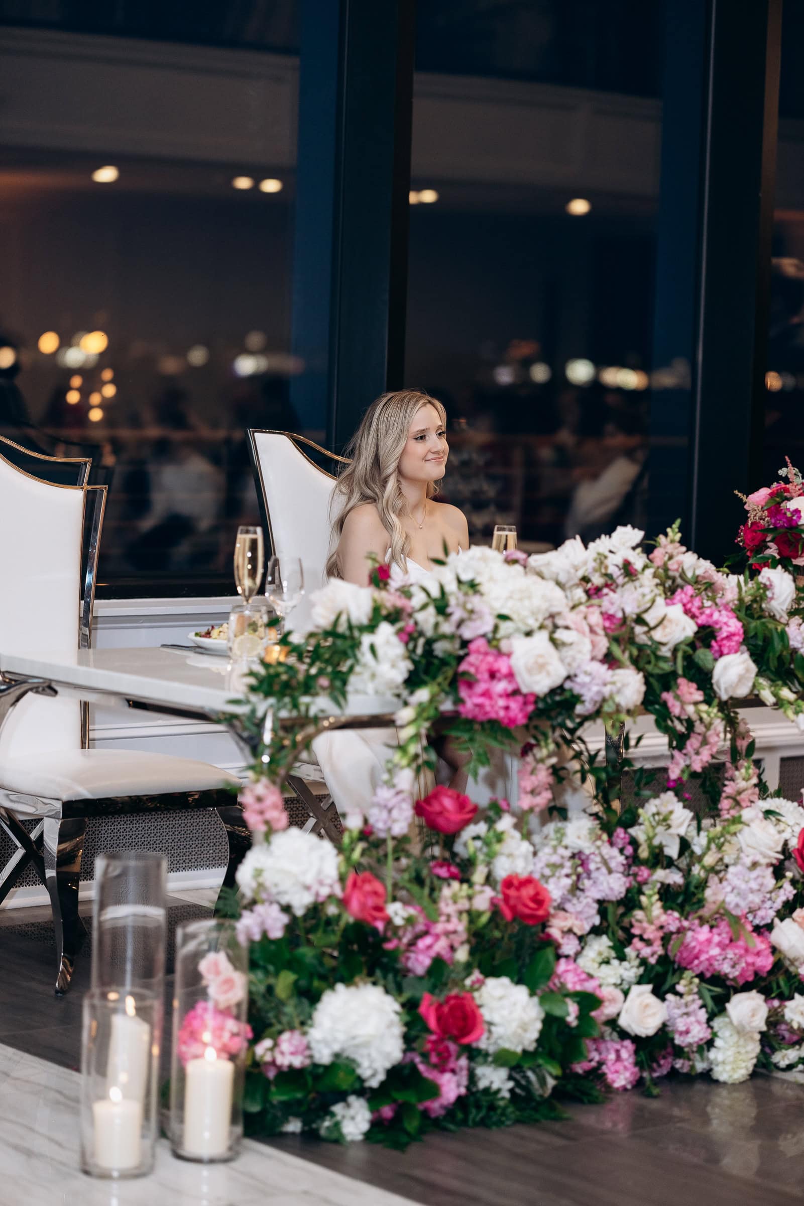 Bride sitting at a white chair behind a lavish arrangement of pink and white flowers and candles at Hudson House, Jersey City, NJ.