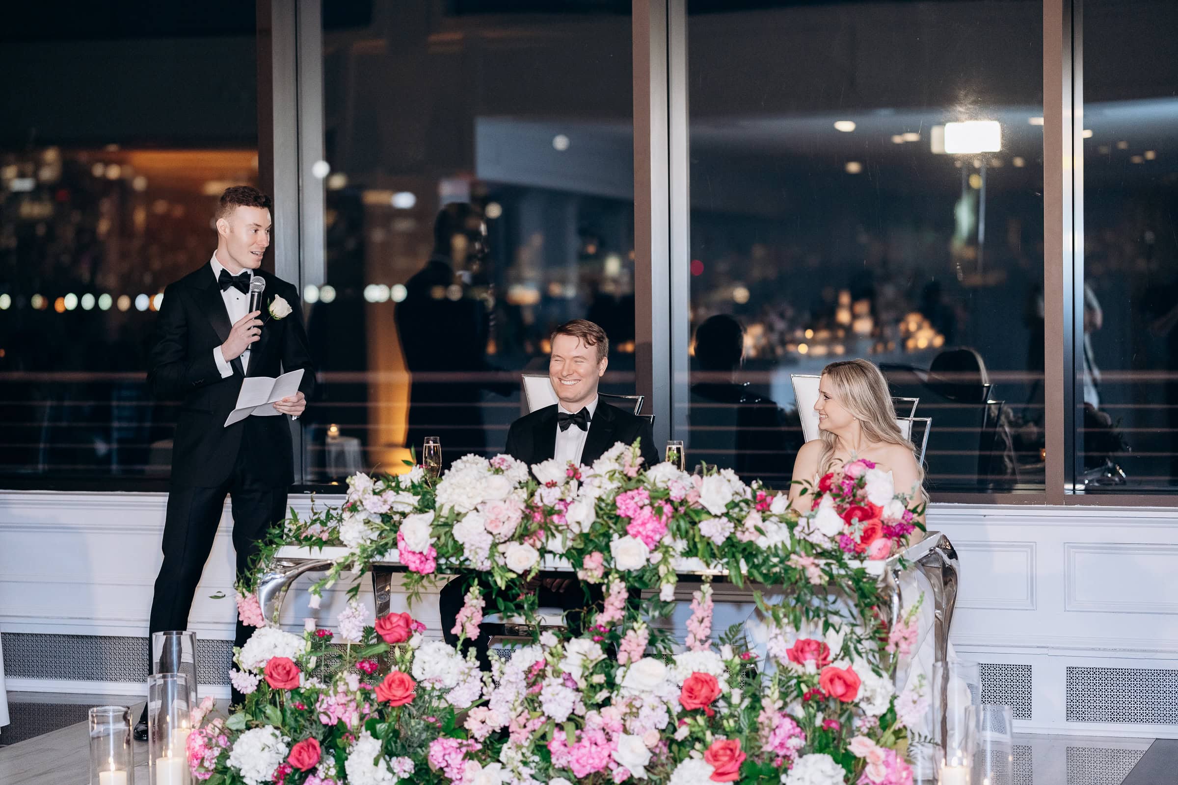 Man in tuxedo giving a speech with microphone while seated couple listens, surrounded by pink and white floral arrangements at Hudson House, Jersey City.