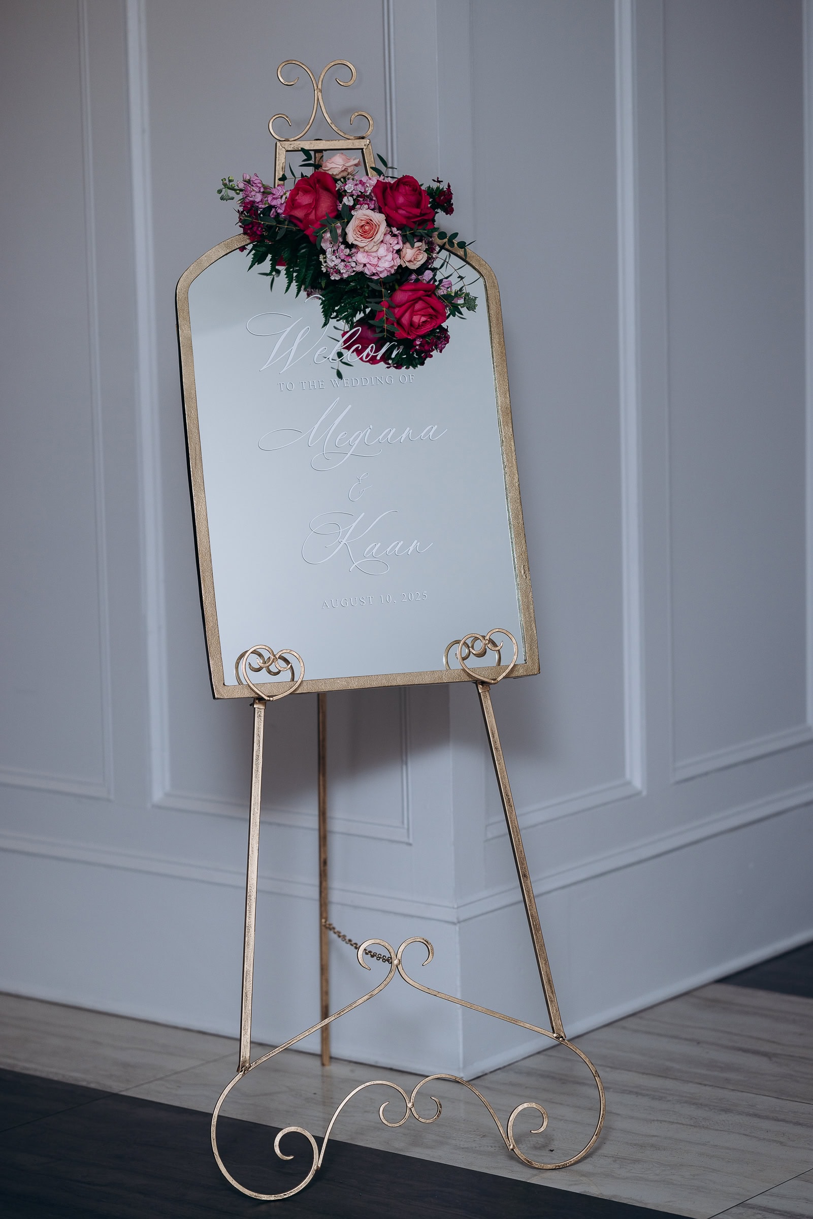 Gold-framed mirror wedding welcome sign with pink and red roses on a decorative gold easel in an elegant interior.