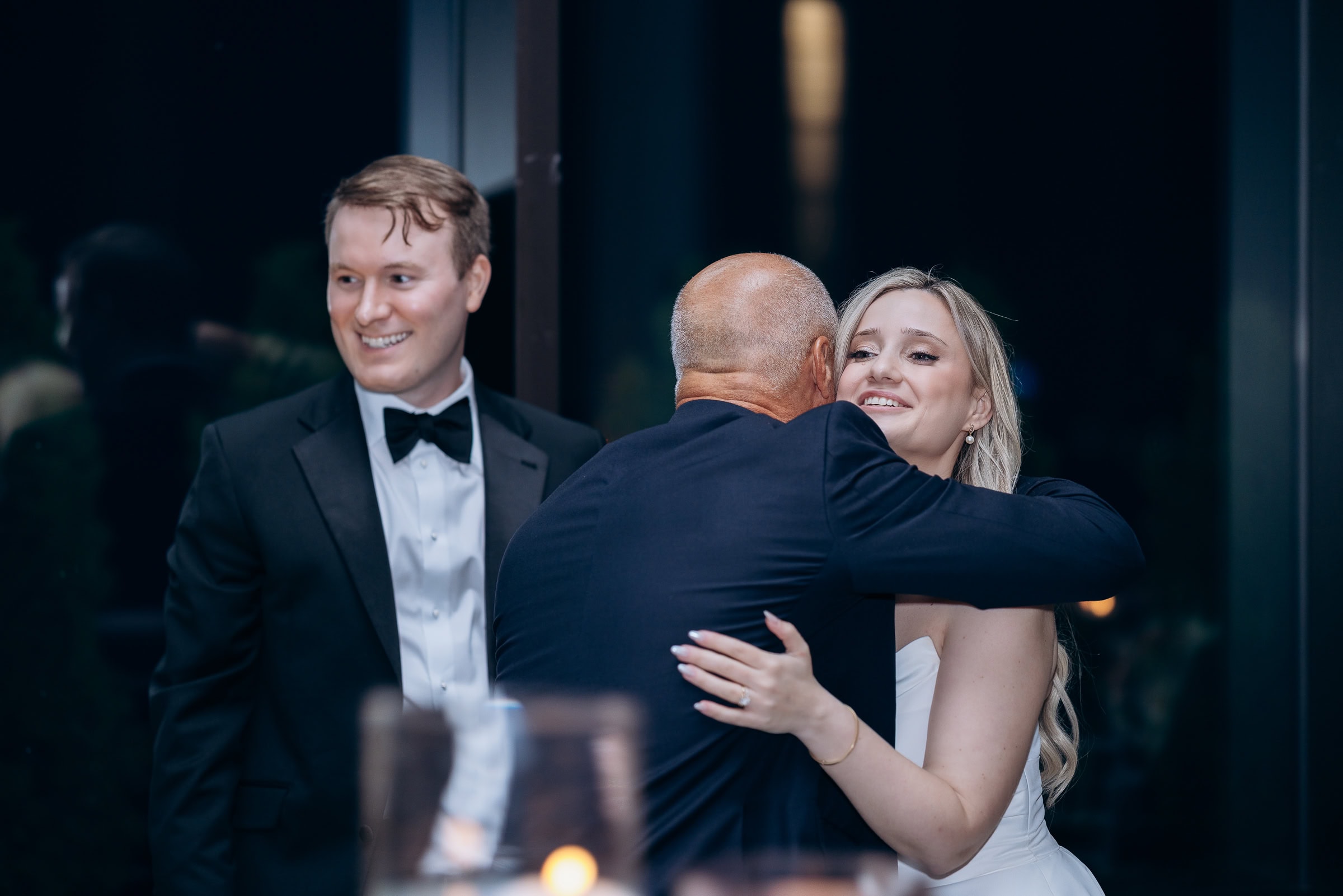 Bride in white gown hugging an older man while a smiling man in a black tuxedo looks on at a Hudson House wedding in Jersey City, NJ.