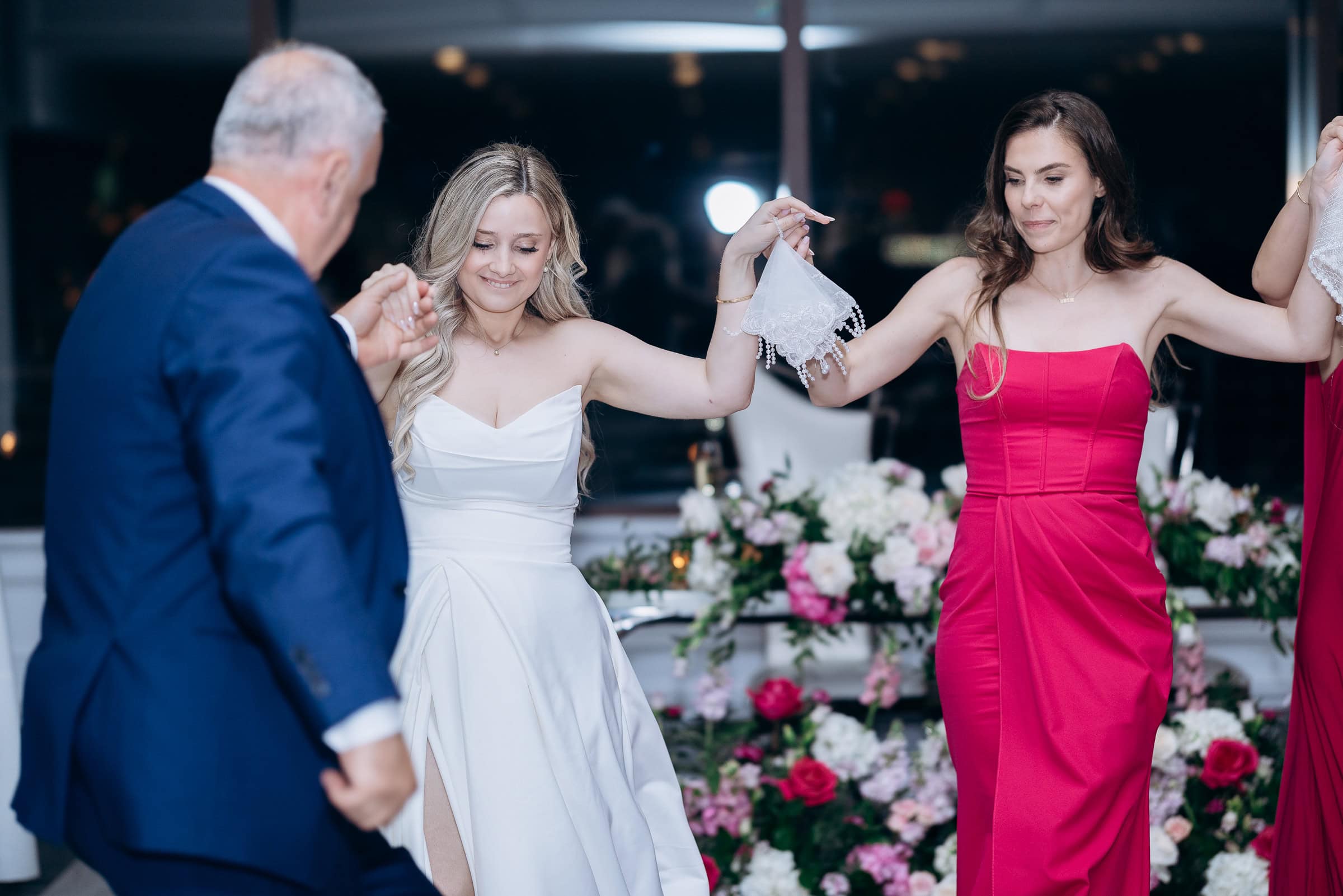 Bride in white gown holding lace handkerchief dancing with woman in crimson dress and man in navy suit at Hudson House wedding.