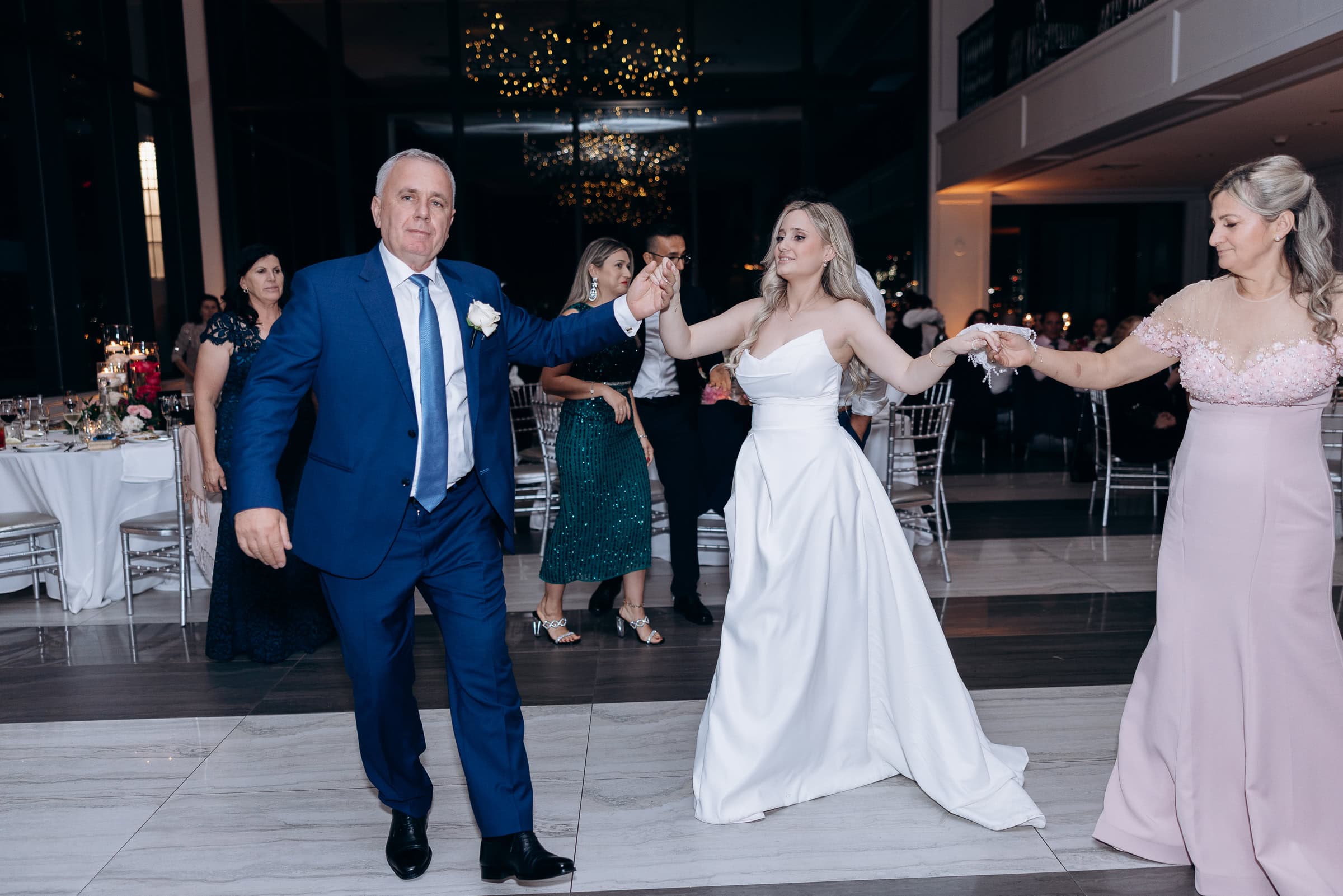 Bride in white gown dances holding hands with two guests in formal attire inside a modern venue with reflective windows and soft chandelier lighting.