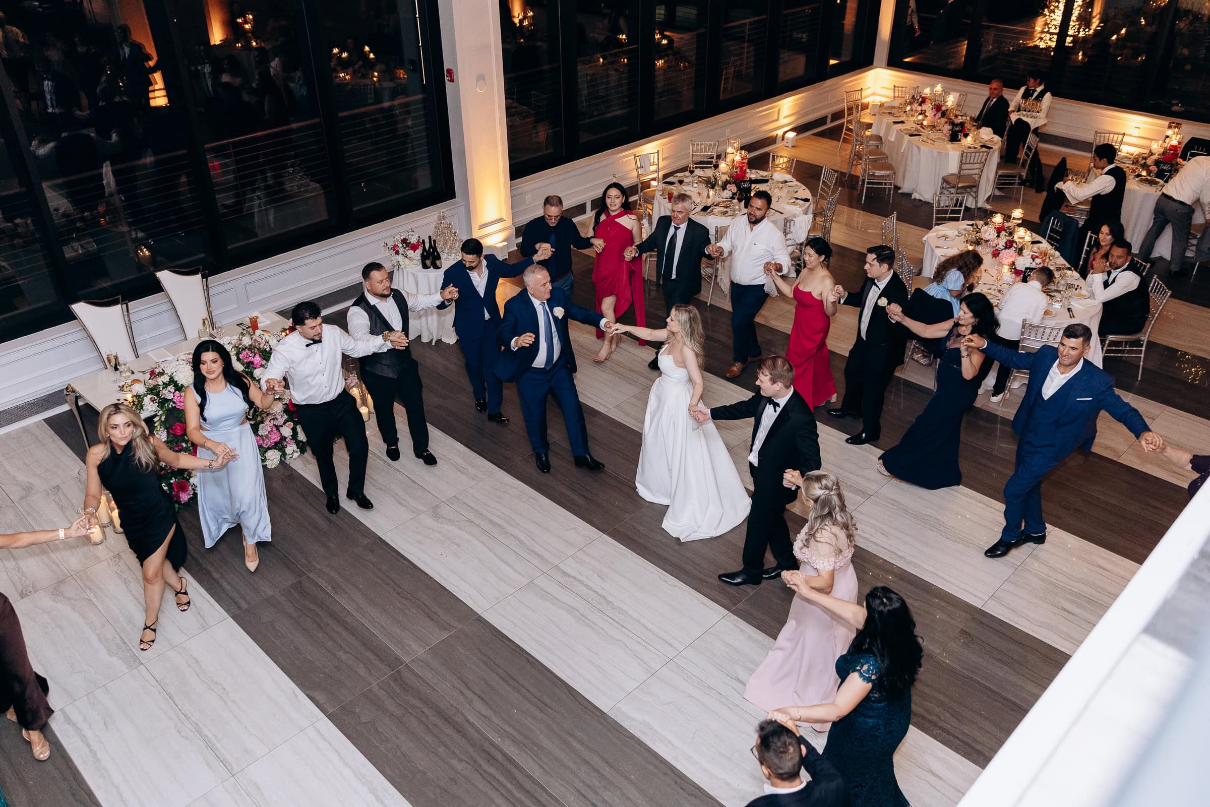 Overhead view of wedding guests holding hands in a circle dance at Hudson House, Jersey City, with candlelit tables and floral arrangements nearby.