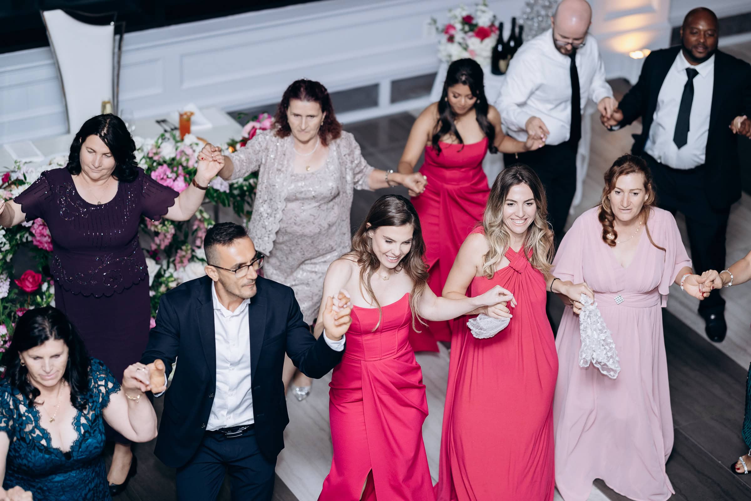 Group of wedding guests holding hands and dancing in a circle indoors, with women in vibrant dresses and men in suits at Hudson House, Jersey City.
