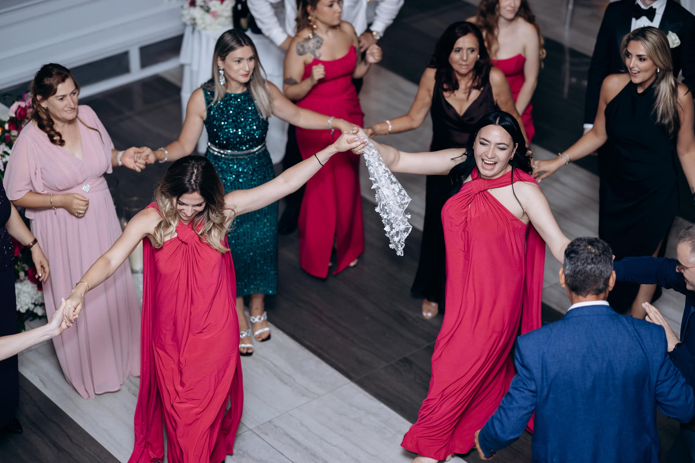 Overhead view of wedding guests holding hands in a dance circle, featuring women in red and green gowns and men in suits at Hudson House, Jersey City, NJ.