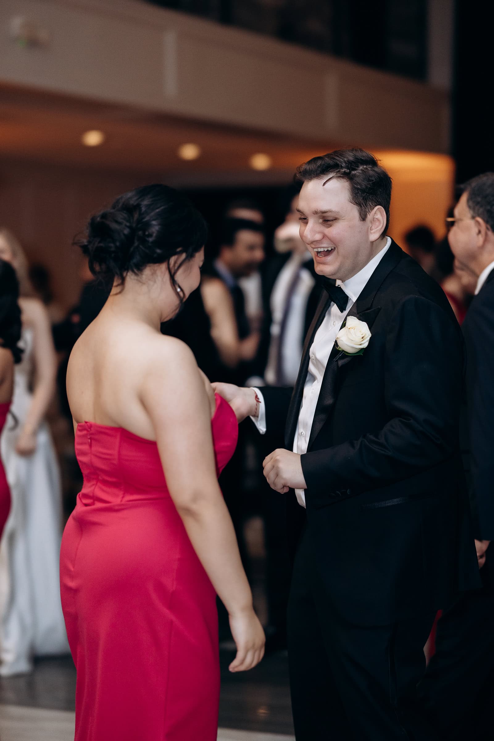 Man in black tuxedo with white rose boutonniere smiling and holding hands with woman in strapless magenta gown at wedding reception.