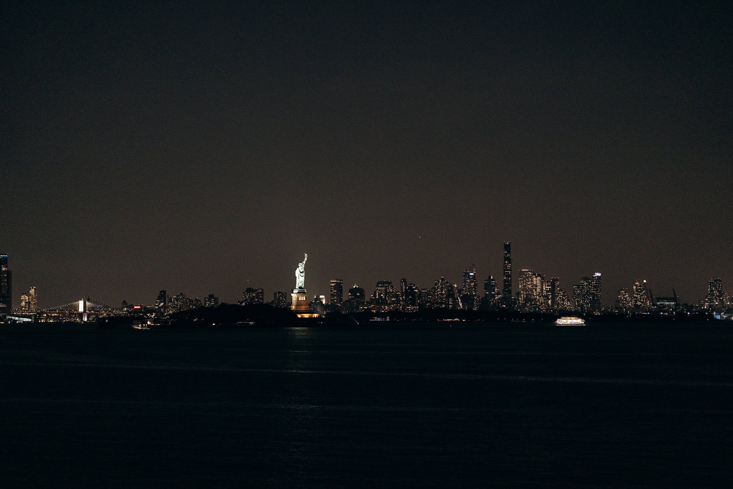 Night view of Manhattan skyline with Statue of Liberty lit up and a boat glowing on the water near Hudson House, Jersey City, NJ.