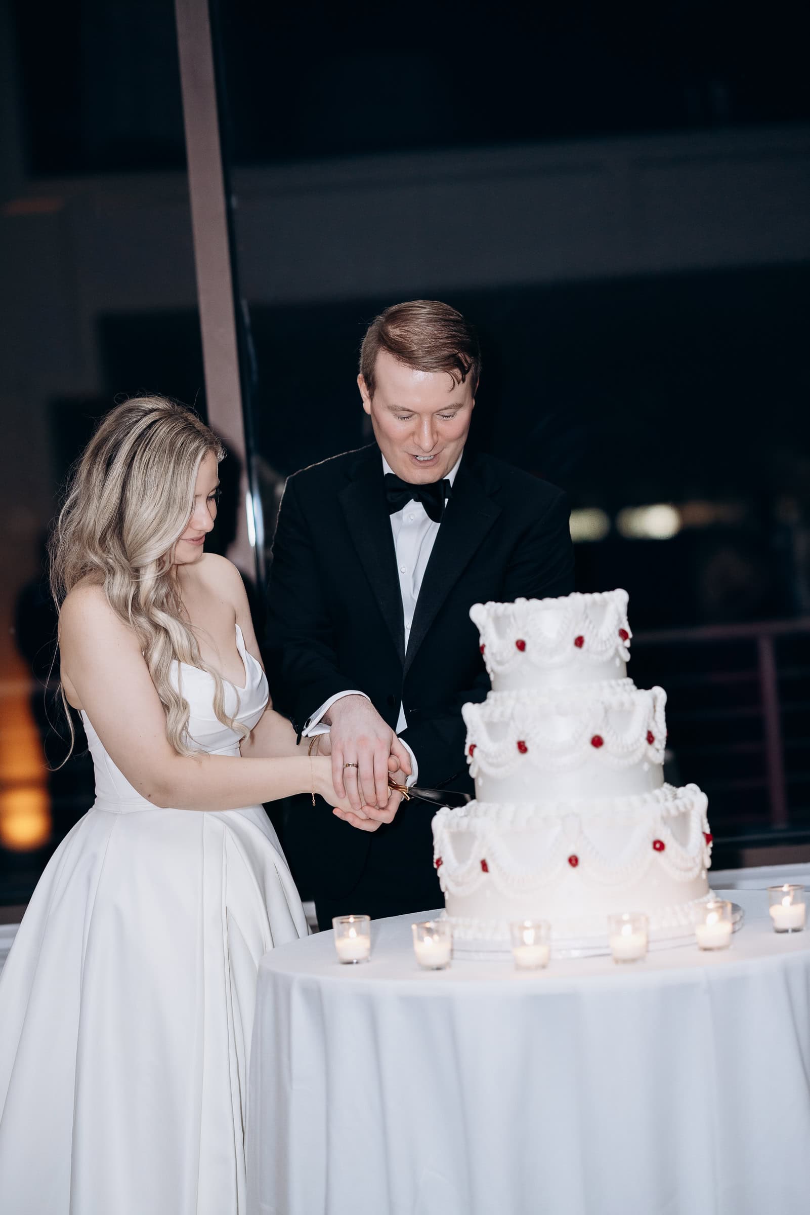 Couple in formal attire cutting a three-tiered white wedding cake with intricate piping and red details, surrounded by candlelight at Hudson House, Jersey City.