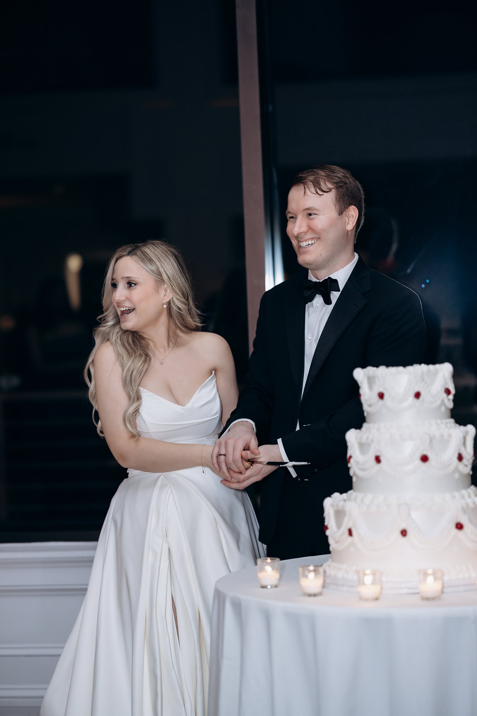 Bride in satin gown and groom in black tuxedo cutting a white tiered wedding cake surrounded by lit candles at Hudson House, Jersey City.