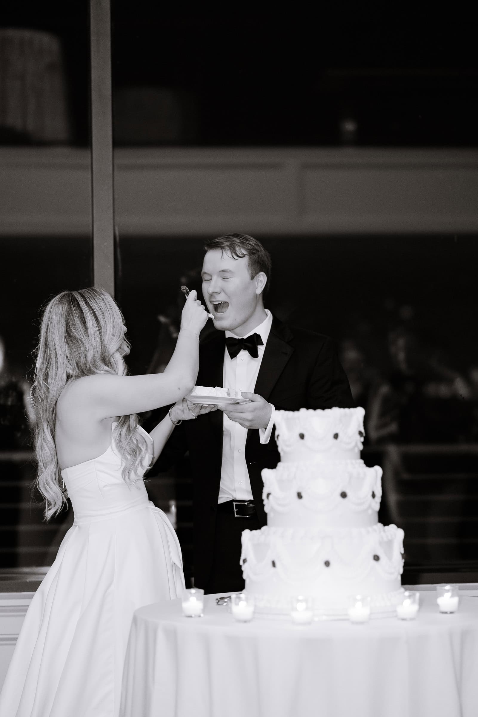 Bride in white satin gown feeding wedding cake to groom in black tuxedo beside tiered cake and candles at Hudson House, Jersey City.