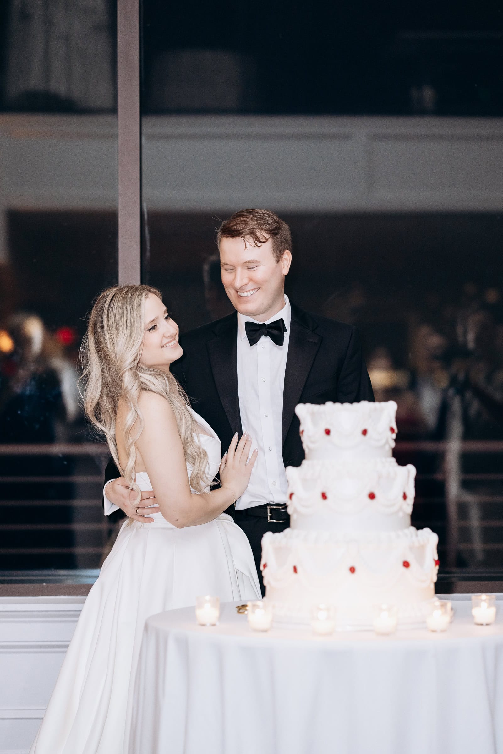 Bride and groom in elegant attire embracing near a four-tiered white wedding cake decorated with scalloped icing and red details, surrounded by lit candles on a table.