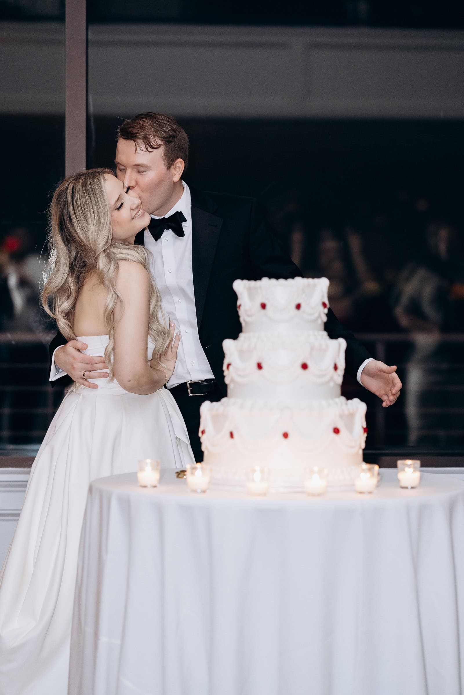 Groom in black tuxedo kisses bride in satin gown near tiered wedding cake with candles at Hudson House, Jersey City.