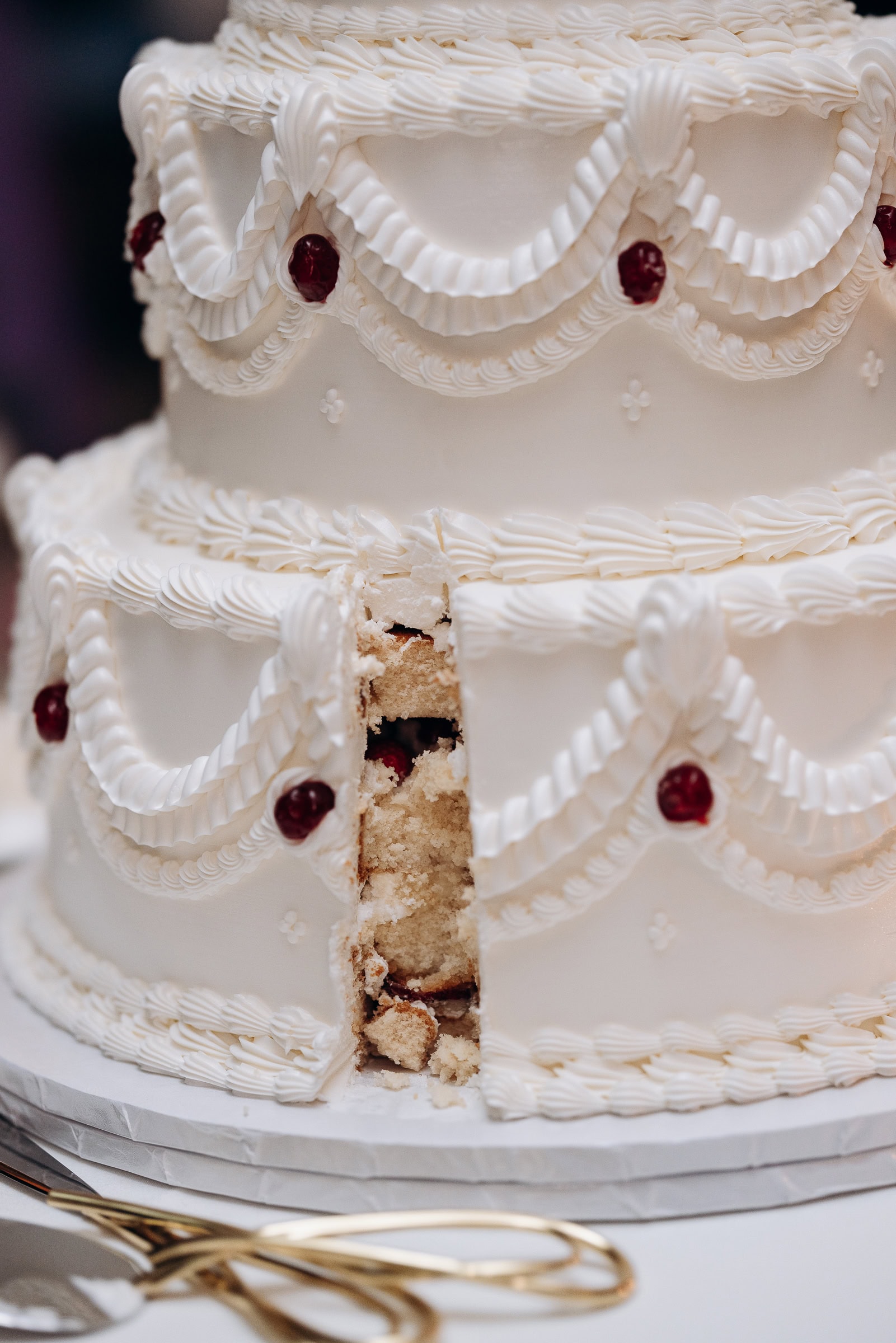 Close-up of a white tiered wedding cake with intricate piped buttercream swags and red berry accents, a slice removed revealing vanilla cake interior.