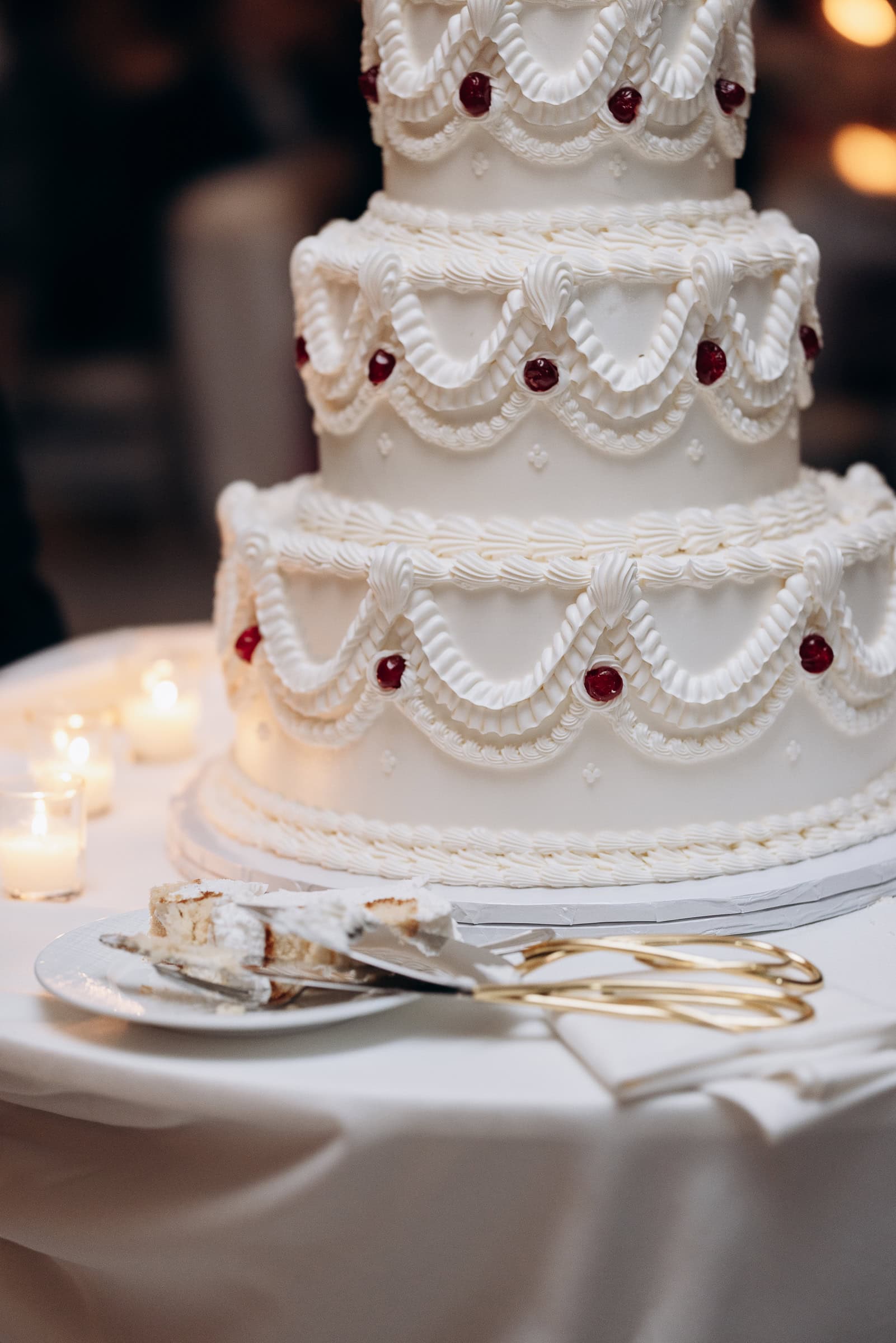 Close-up of a three-tier white wedding cake with detailed piped icing and red cherries, candles softly glowing in the background at Hudson House, Jersey City.