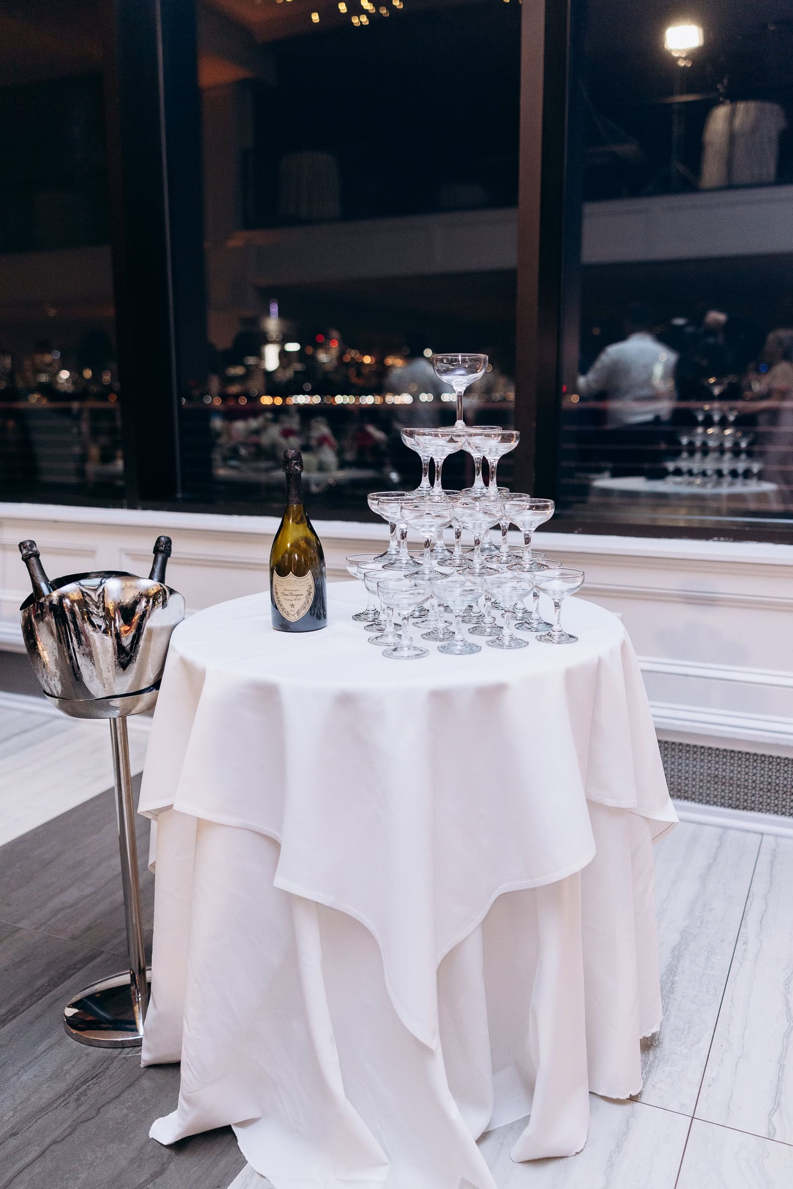 Stacked coupe champagne glasses on a white linen-covered table with a Dom Pérignon bottle and metal ice bucket, Hudson House, Jersey City at night.
