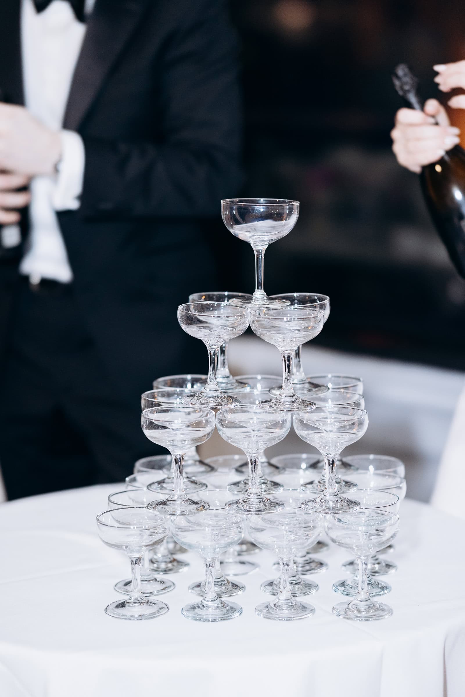 Stacked coupe champagne glasses forming a pyramid on a white tablecloth, with a person in a tuxedo and another holding a champagne bottle blurred in the background at Hudson House, Jersey City.
