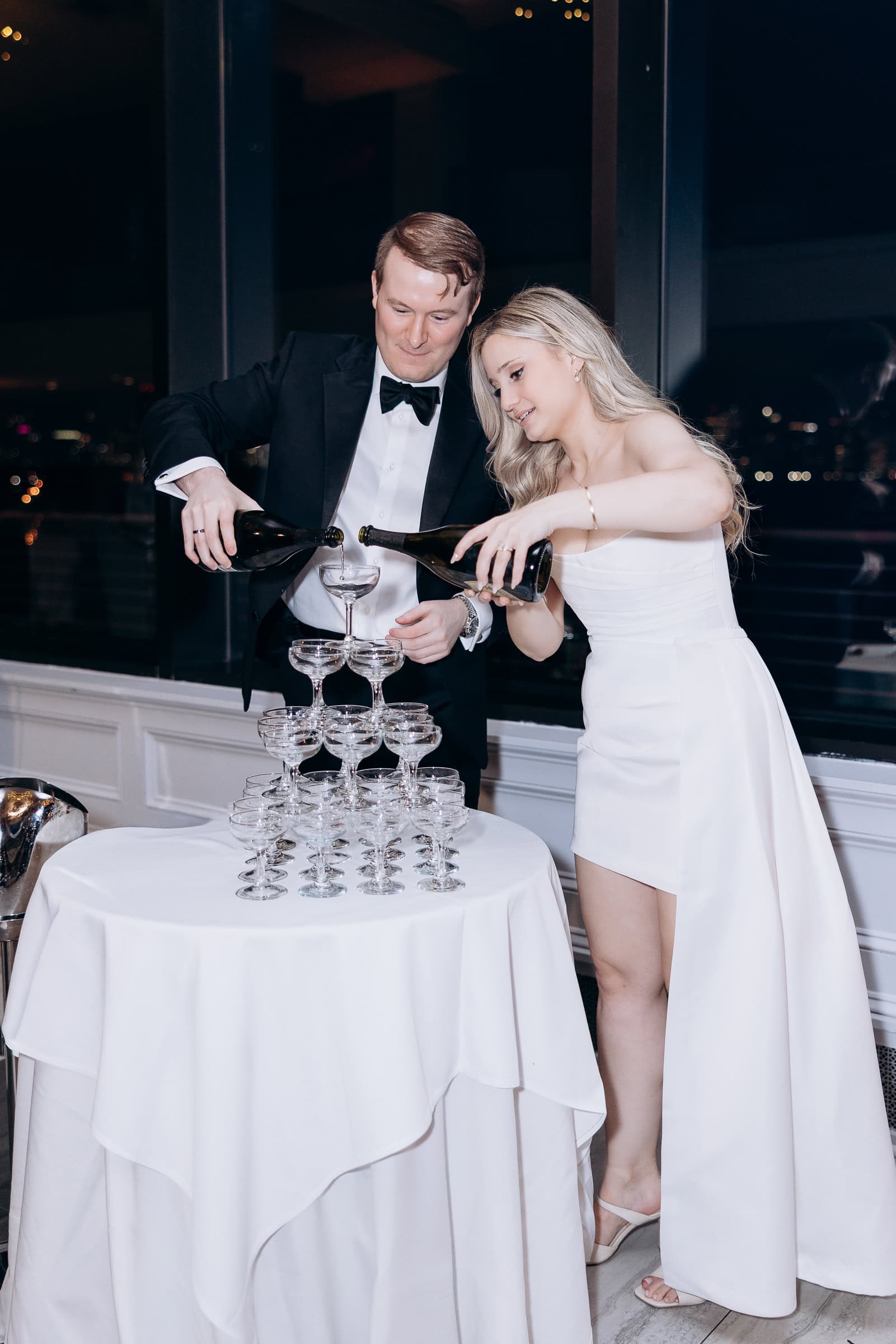 Couple in formal wear pouring champagne into a tower of coupe glasses at Hudson House, Jersey City, NJ, during evening celebration.