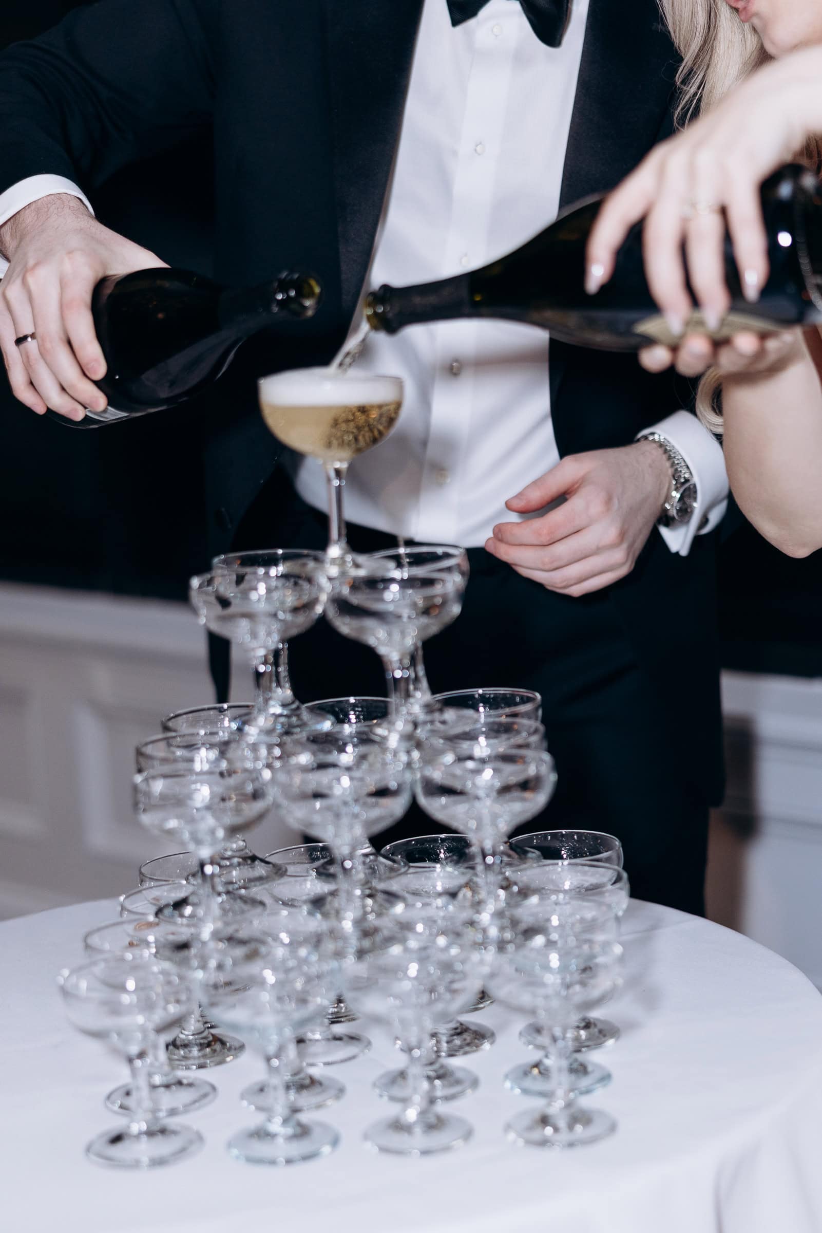 Close-up of two people in formal wear pouring champagne into the top glass of a multi-tiered champagne tower on a white tablecloth.