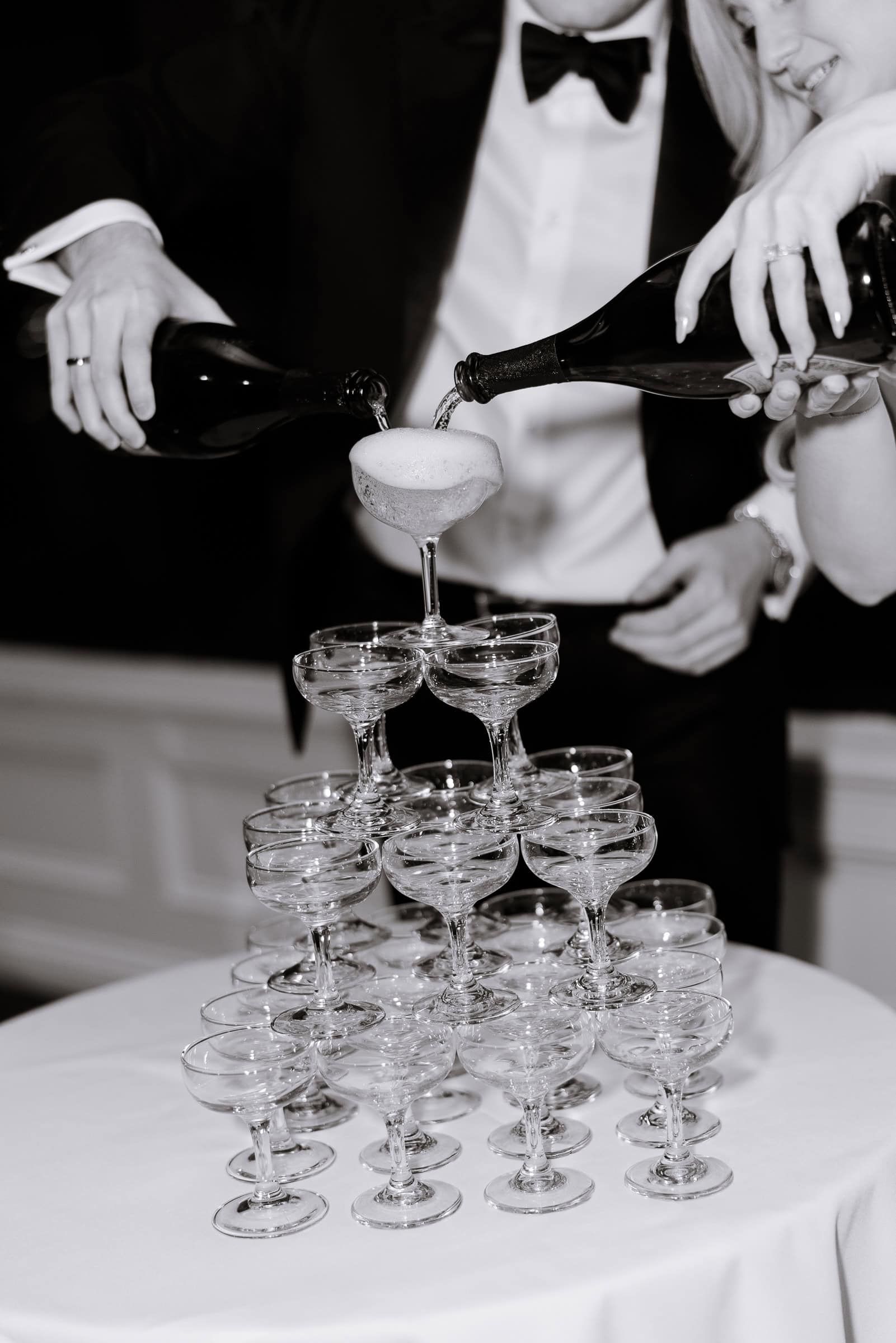 Close-up of a couple pouring champagne from two bottles into the top glass of a pyramid of coupe glasses, with the man in a tuxedo and the woman wearing a ring.