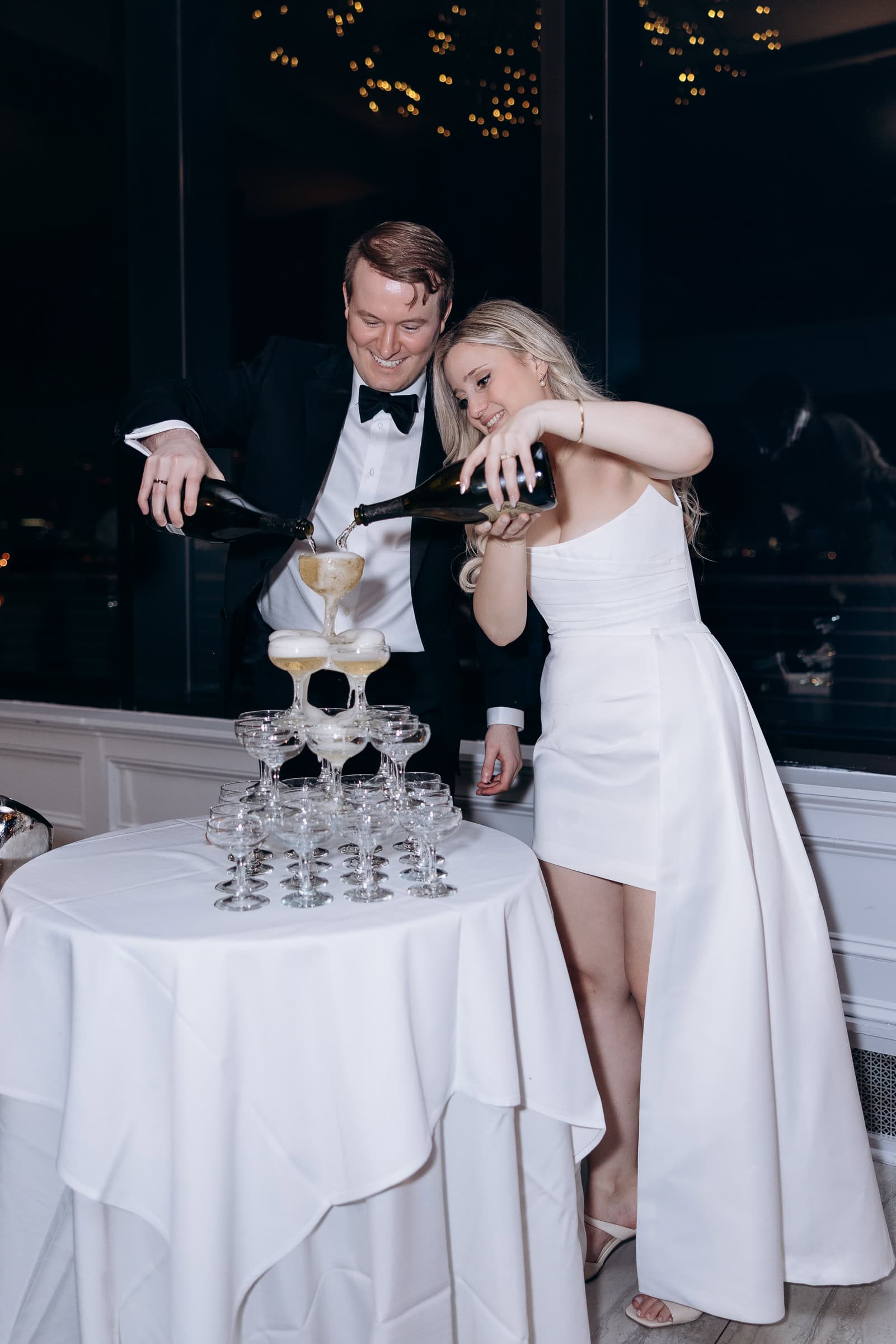 Couple in formal attire pouring champagne into a multi-tiered tower of coupe glasses at Hudson House, Jersey City, NJ, with city lights reflected in window.