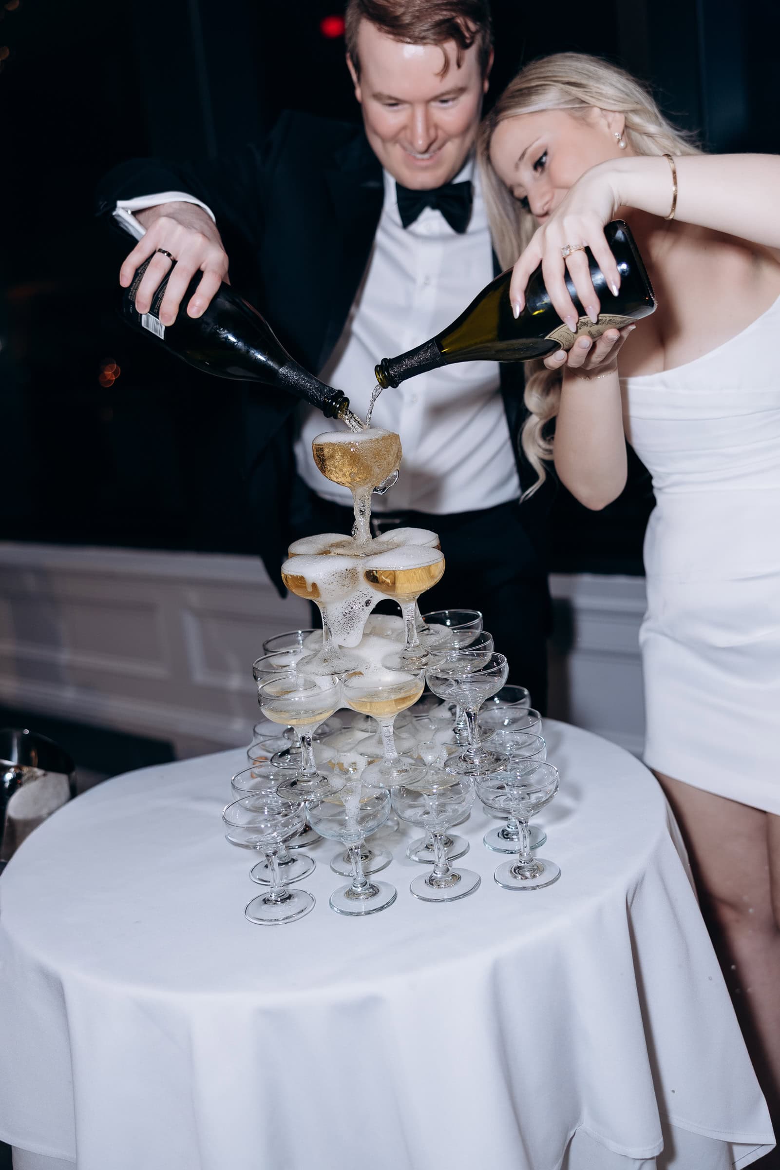 Man and woman in formal attire pouring champagne from two bottles into a stacked tower of coupe glasses on a white tablecloth at Hudson House, Jersey City.