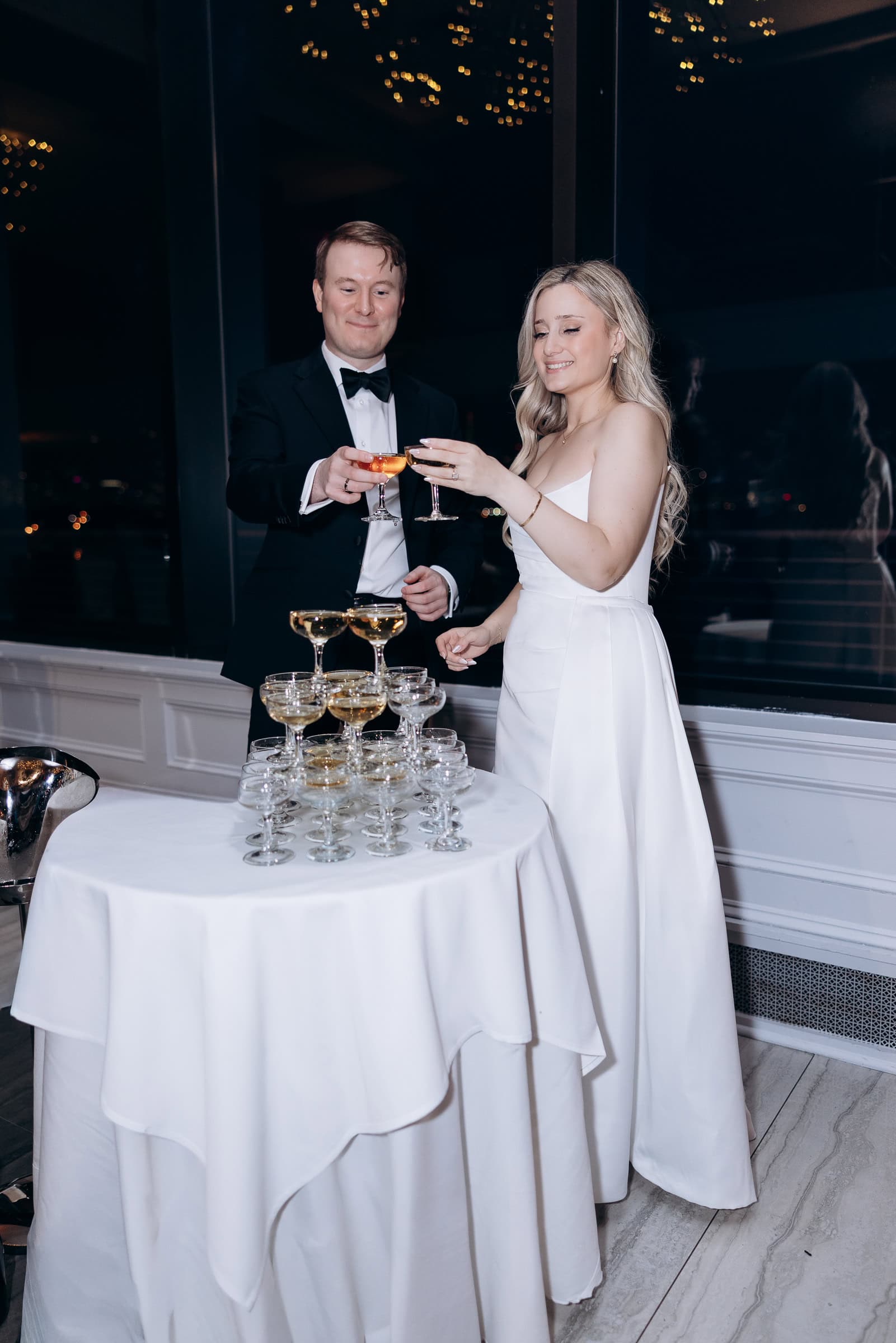 Couple in formal attire toasting champagne glasses next to a tower of champagne coupes at Hudson House, Jersey City, NJ.