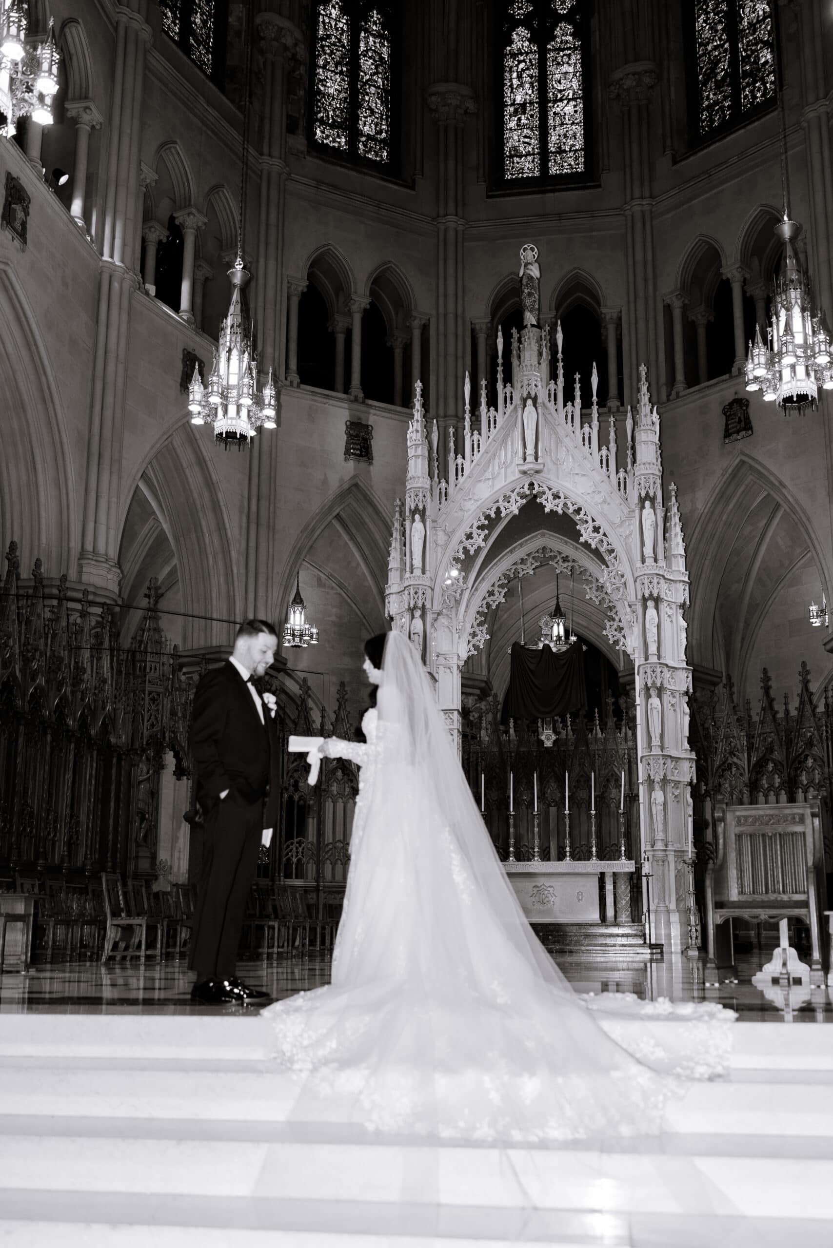 Black and white photo of a bride and groom standing on marble steps inside The Cathedral Basilica of the Sacred Heart, framed by gothic arches and illuminated chandeliers.