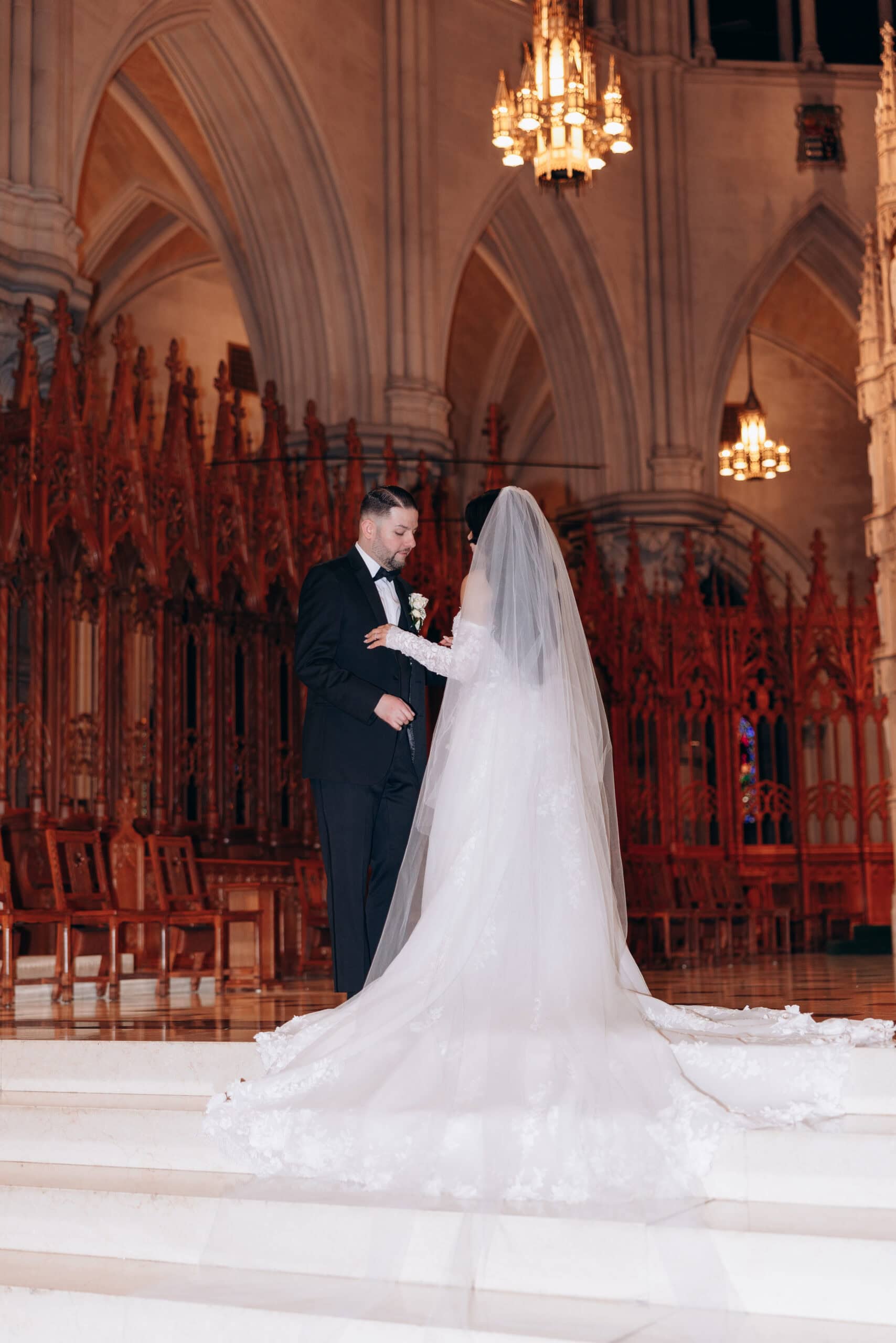 Bride and groom standing on marble steps inside The Cathedral Basilica of the Sacred Heart, surrounded by Gothic arches and intricate wood carvings, illuminated by warm chandeliers.