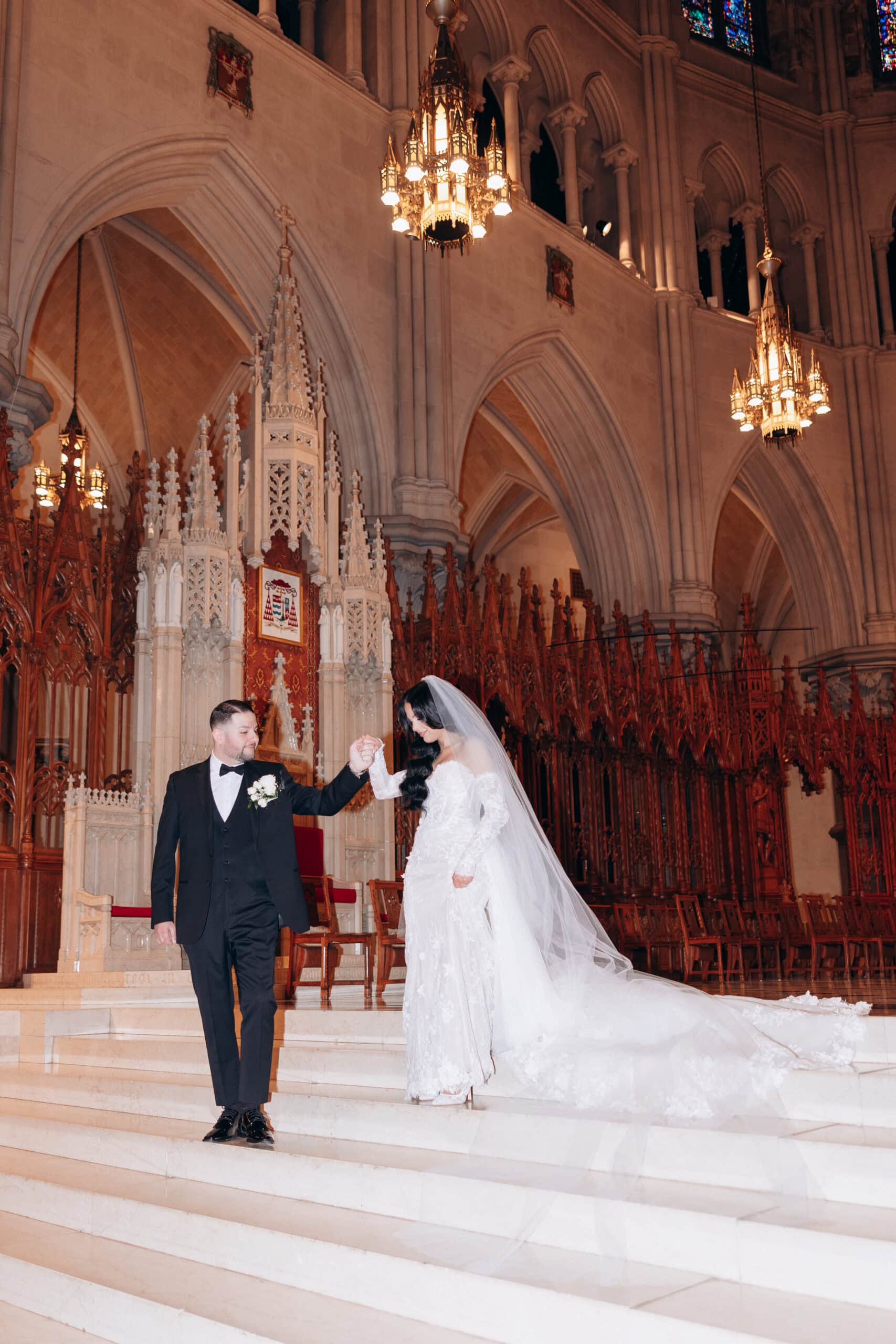 Groom in black tuxedo holding bride's hand as she descends marble steps inside The Cathedral Basilica of the Sacred Heart, surrounded by gothic arches and chandeliers.