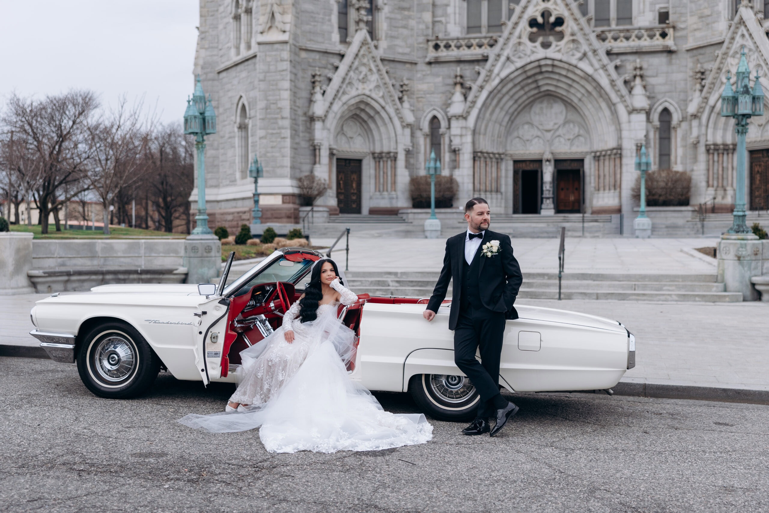 Bride in lace wedding dress sitting in a white vintage Thunderbird with red interior, groom in black tuxedo leaning on car, in front of The Cathedral Basilica of the Sacred Heart.