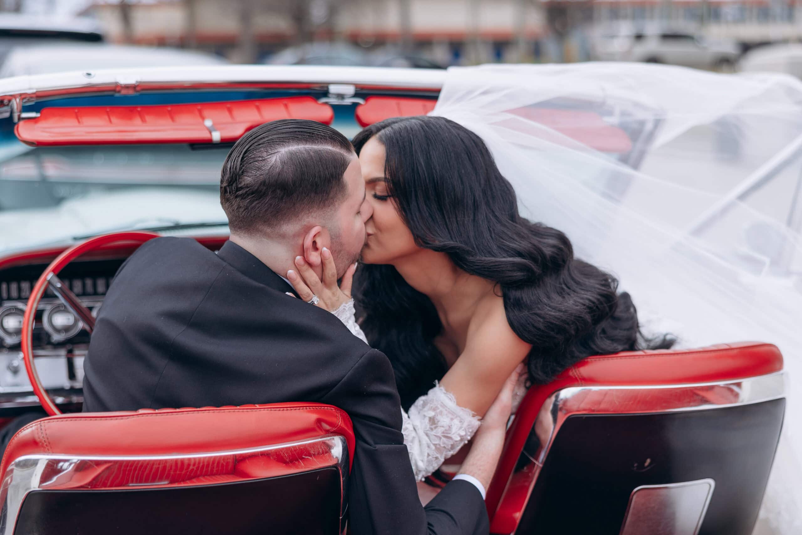 Bride and groom kissing inside a vintage convertible with red leather seats, bride's veil flowing, captured in an intimate moment.