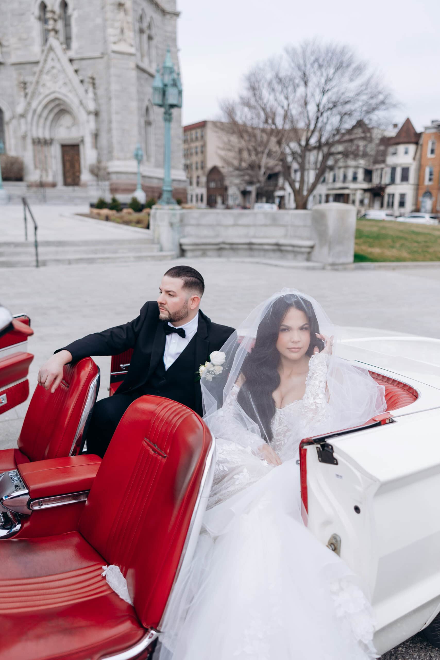 Bride in lace wedding gown and veil with groom in black tuxedo seated in white vintage convertible with red leather interior outside Cathedral Basilica of the Sacred Heart.