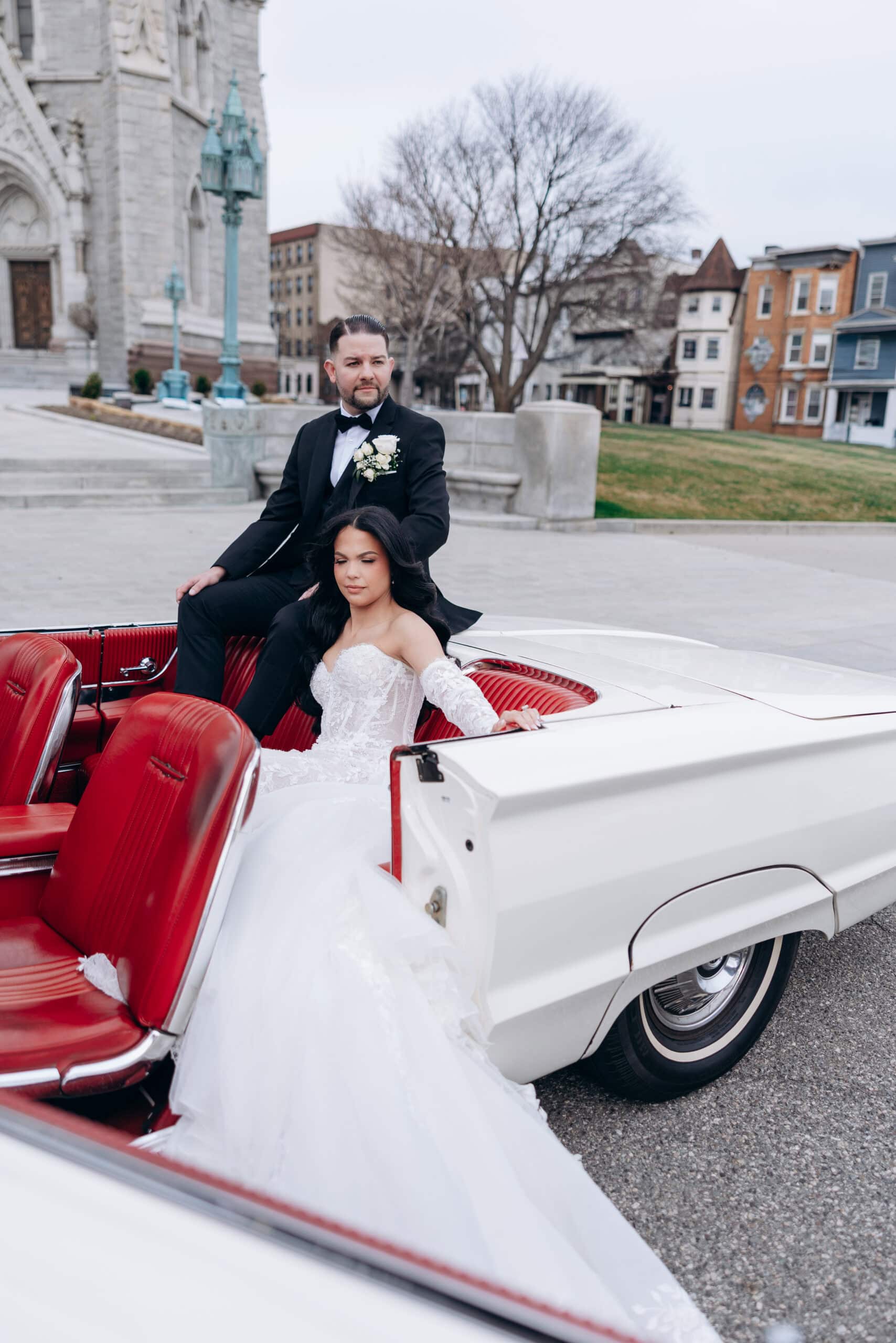 Bride in lace wedding dress seated in a vintage white convertible with red leather interior, groom in black tuxedo standing beside her, historic cathedral facade in background.