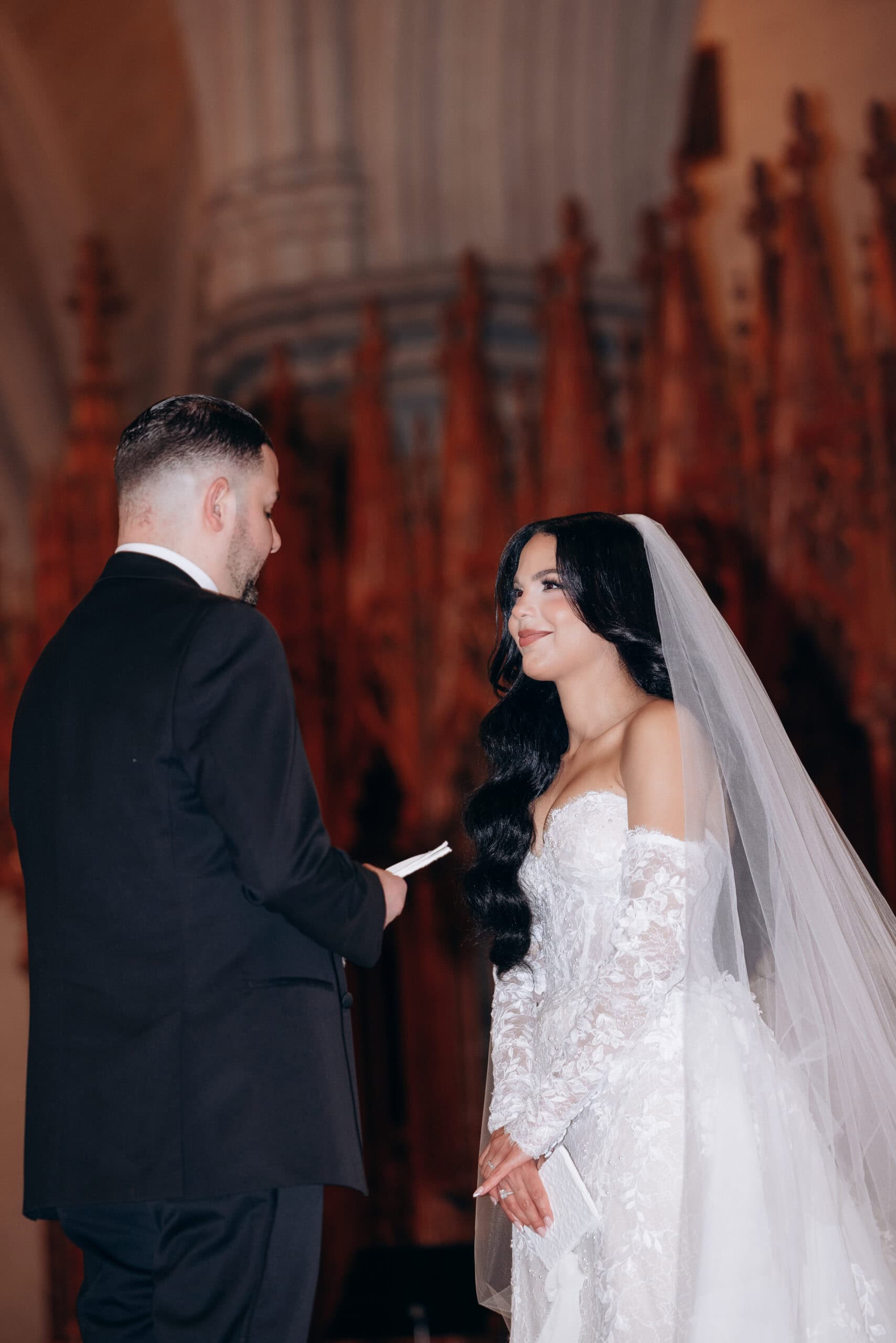 Bride in lace wedding dress and veil holding a white handkerchief, facing groom in black suit, exchanging vows inside The Cathedral Basilica of the Sacred Heart with detailed wooden carvings in background.