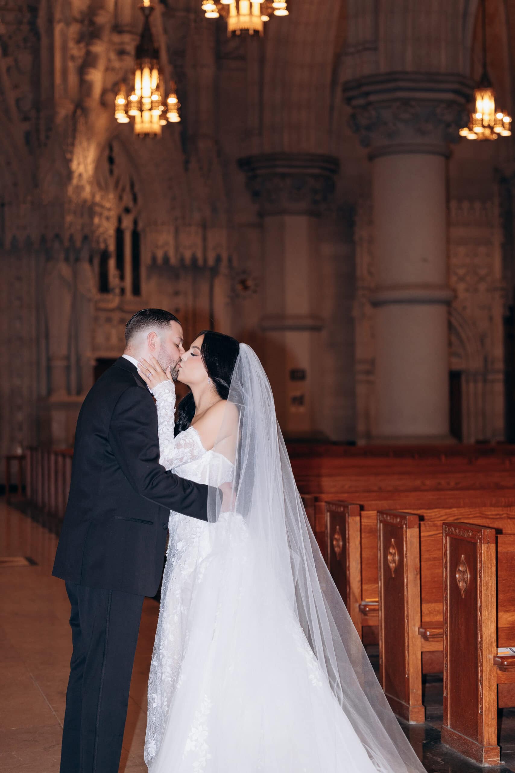 Bride and groom kissing inside The Cathedral Basilica of the Sacred Heart, surrounded by gothic arches, detailed stonework, and warm chandelier lighting.