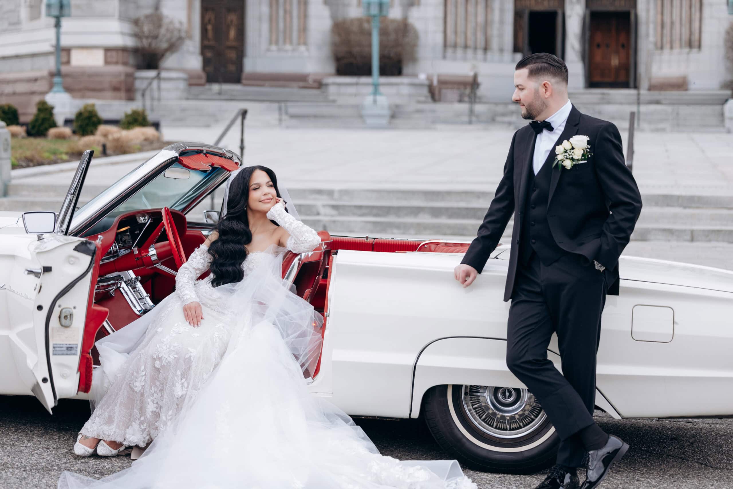 Bride in lace wedding gown seated in vintage white convertible with red interior, groom in black tuxedo leaning on car, steps of The Cathedral Basilica of the Sacred Heart in background.