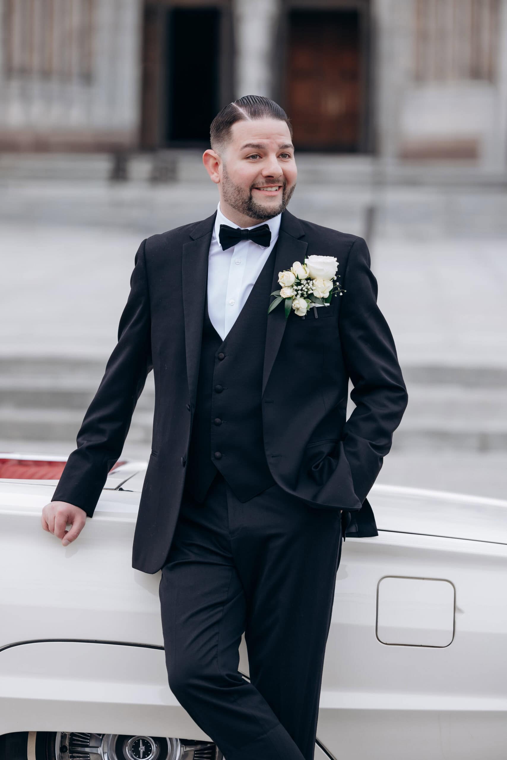 Man in black tuxedo with white rose boutonniere leaning on a white vintage car, in front of stone steps and large wooden doors at The Cathedral Basilica of the Sacred Heart.