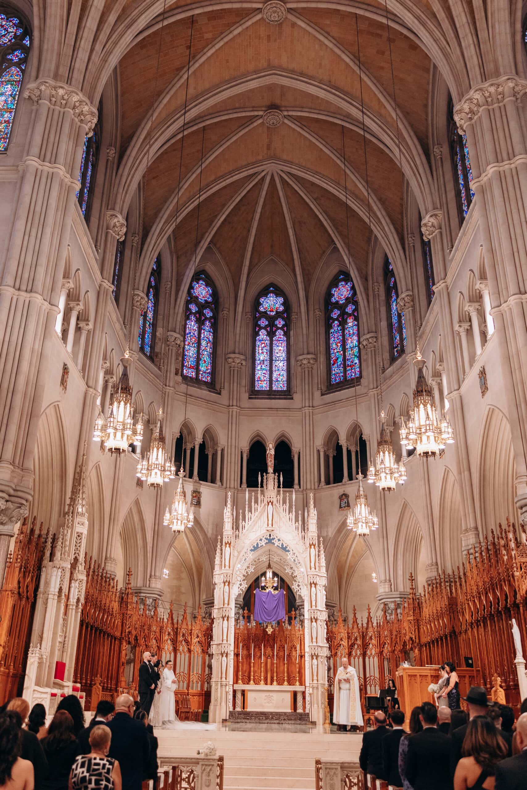 Interior view of The Cathedral Basilica of the Sacred Heart during a wedding ceremony, showcasing vaulted ceilings, stained glass windows, chandeliers, and guests seated facing the altar.