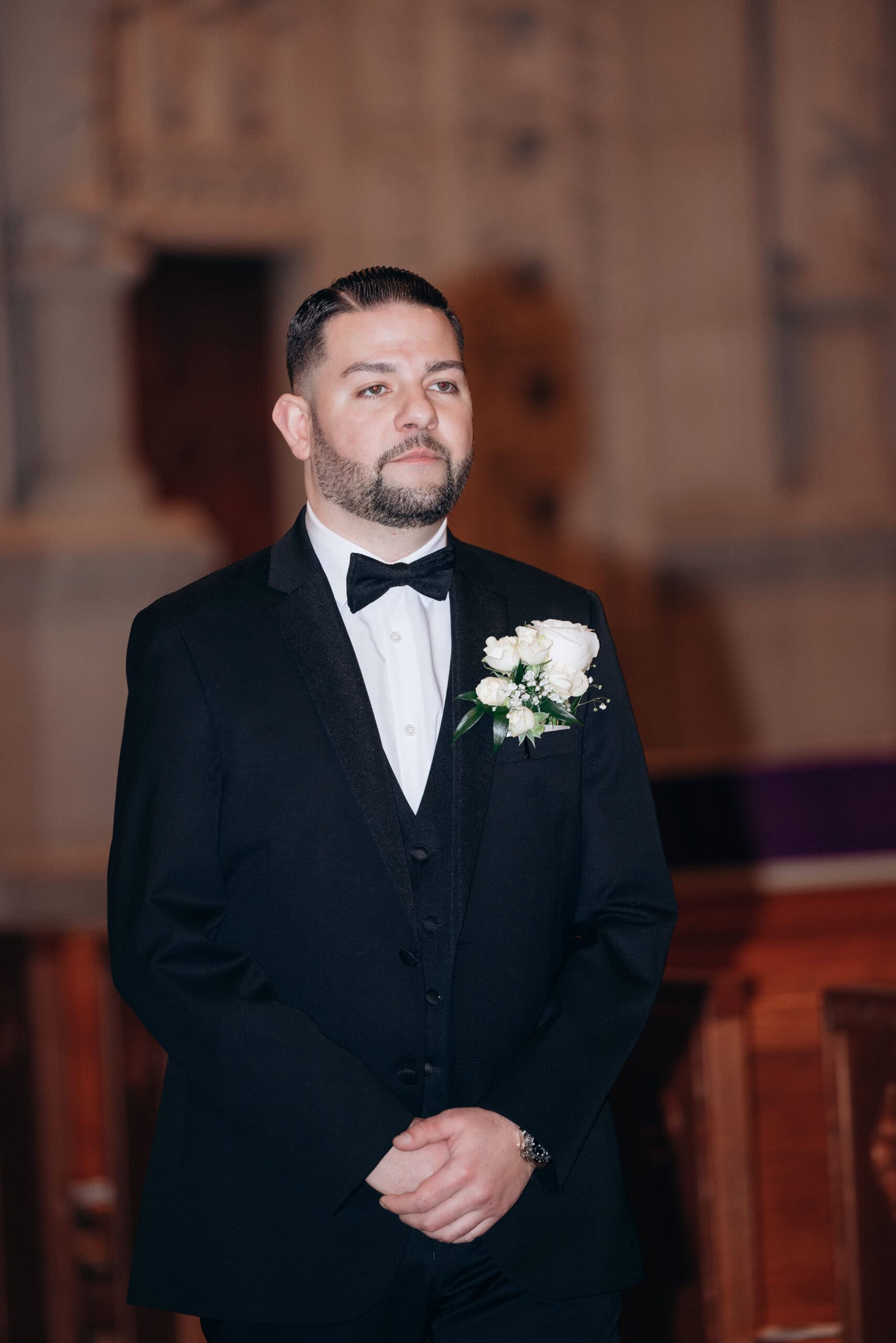 Man in black tuxedo with bow tie and white rose boutonniere standing inside a church with blurred architectural details in the background.