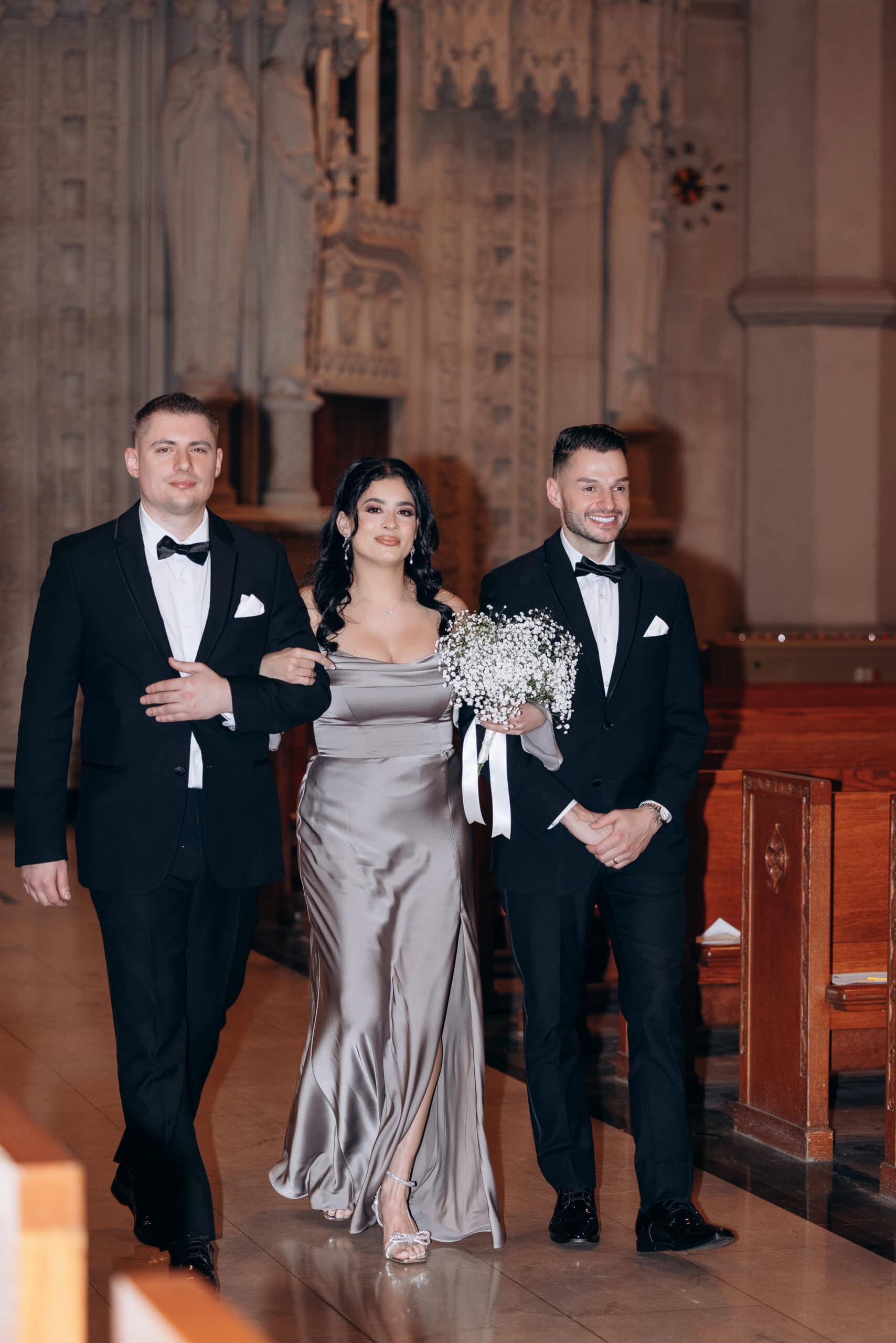 Woman in satin taupe gown holding baby's breath bouquet, flanked by two men in black tuxedos inside The Cathedral Basilica of the Sacred Heart.