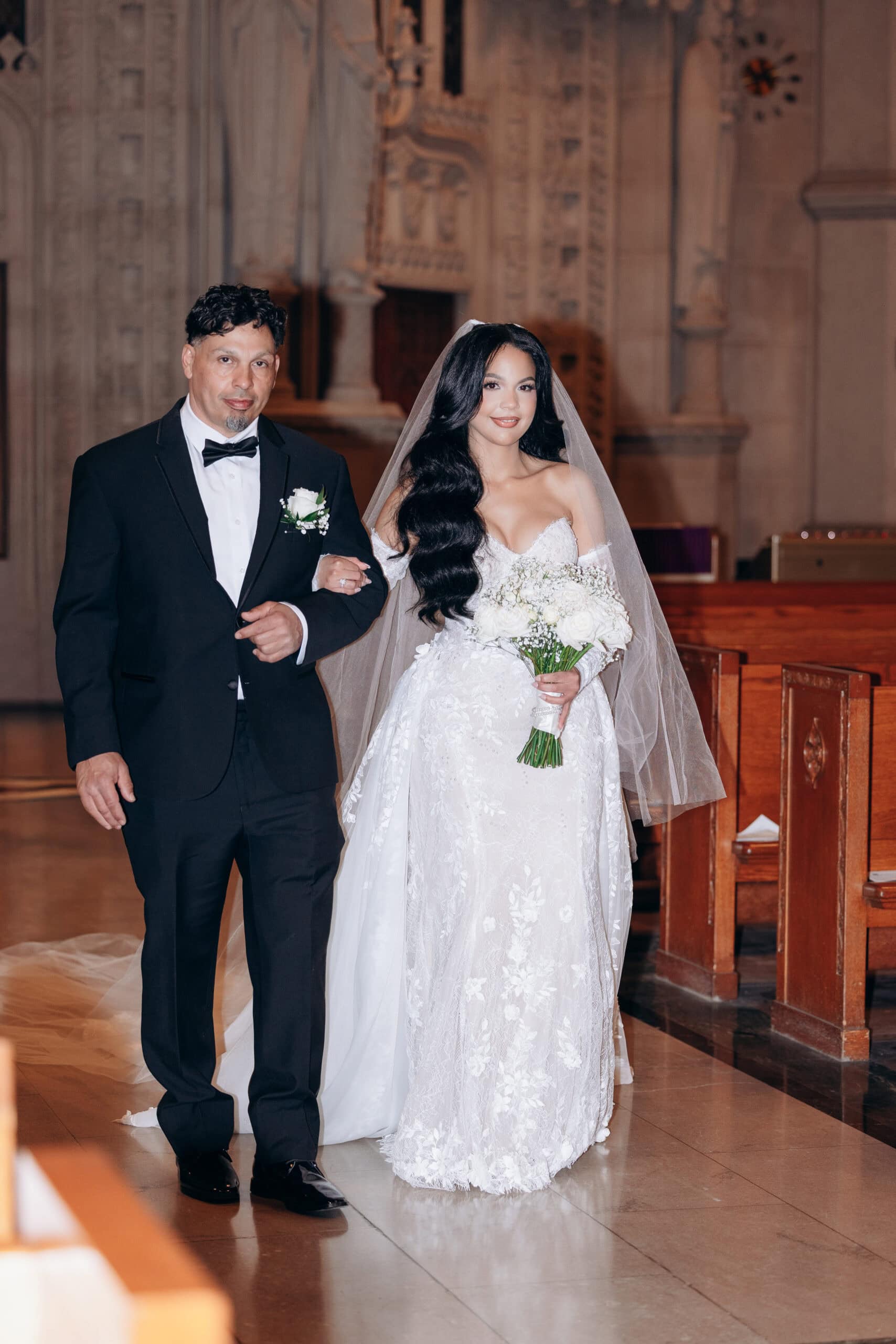 Bride in lace off-shoulder wedding gown with veil holding white rose bouquet walking arm-in-arm with man in black tuxedo inside The Cathedral Basilica of the Sacred Heart.